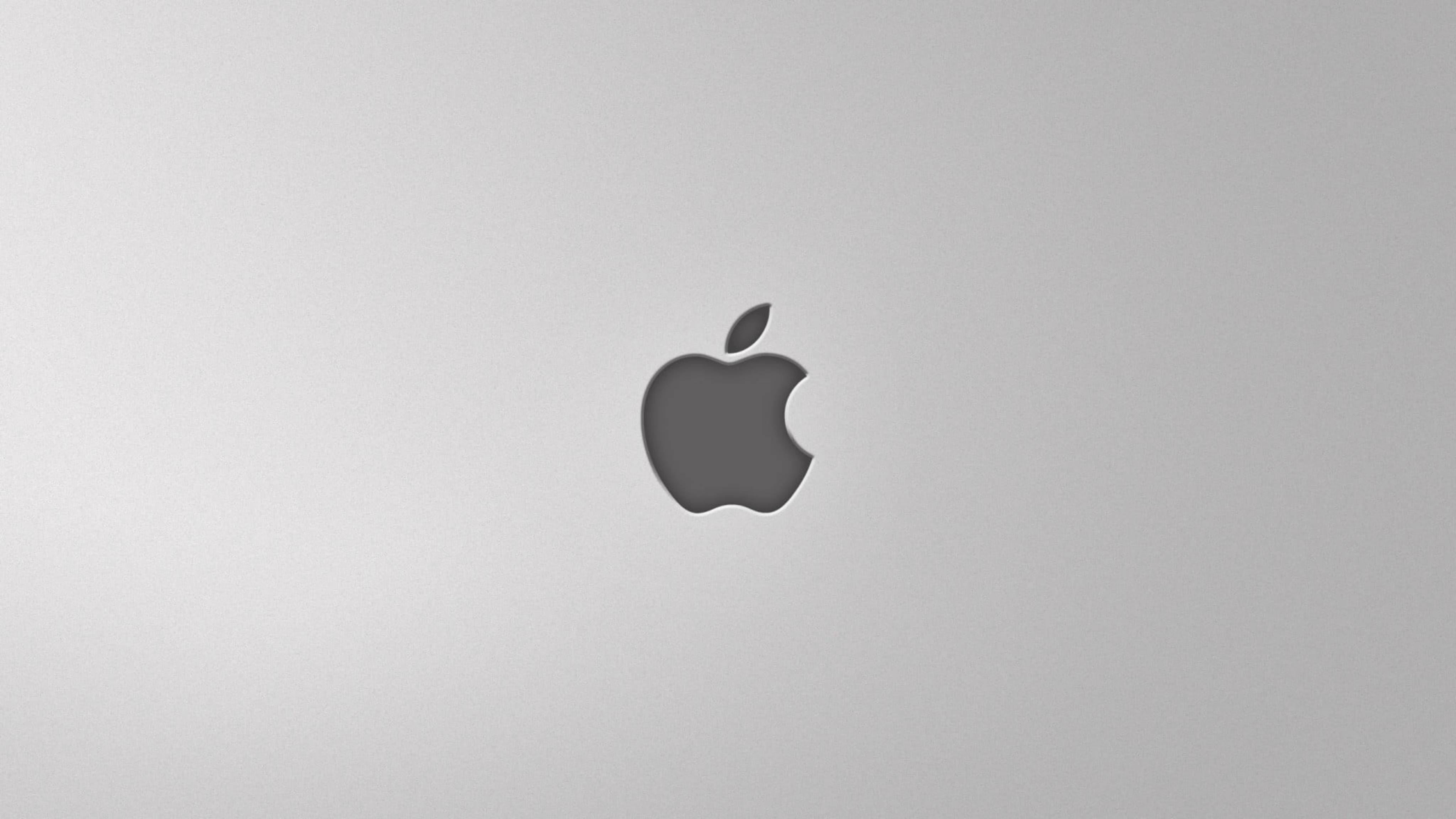 Apple 4K Ultra HD 桌布 壁紙 — 一個簡約的灰色Apple標誌，置中於拉絲金屬筆記型電腦蓋上，帶有柔和光線。