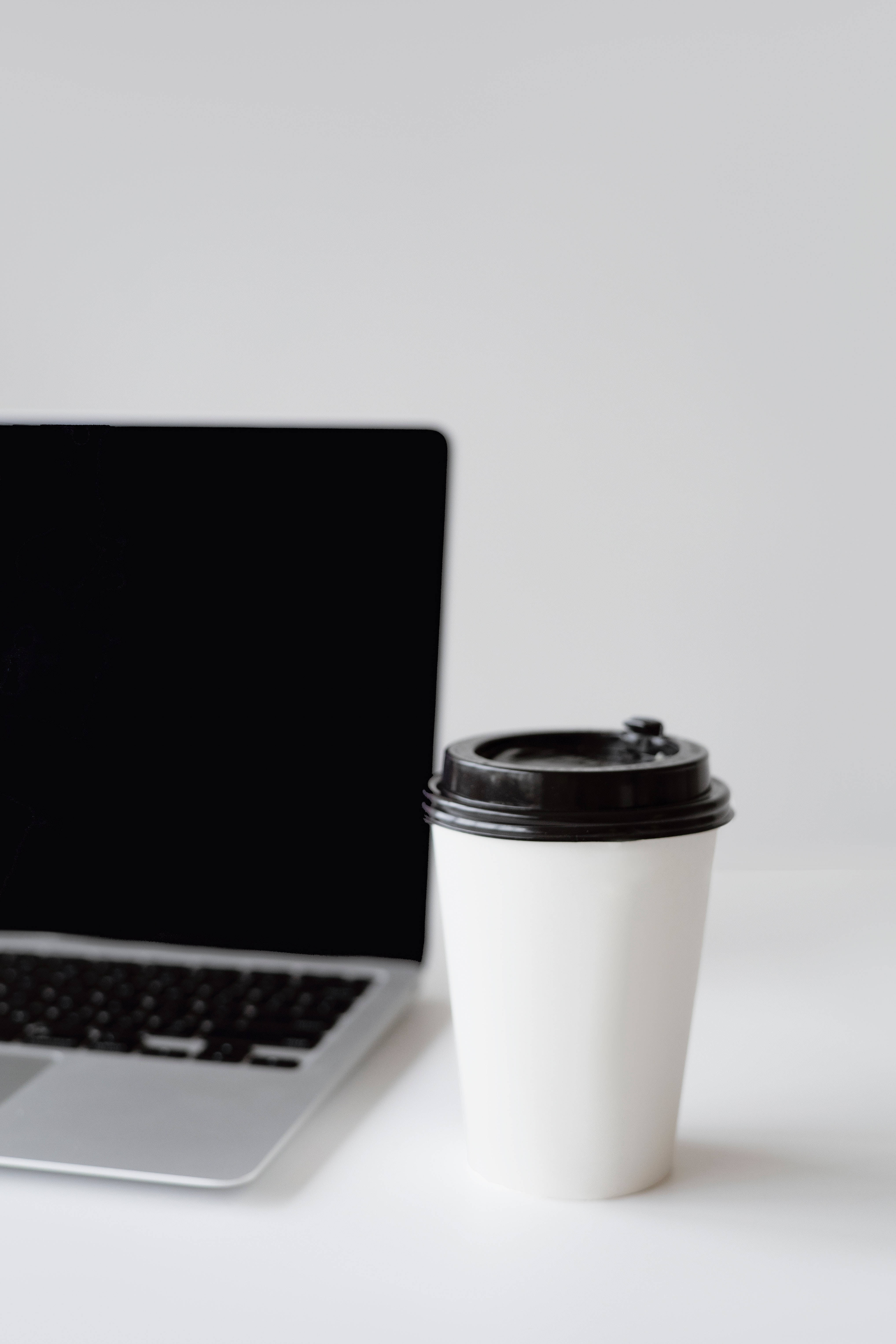 aesthetic tumblr laptop 桌布 壁紙 — 一個簡約的白色咖啡杯（附黑色蓋）放在一個乾淨的白色表面上，旁邊是一部關閉的銀色筆記型電腦。