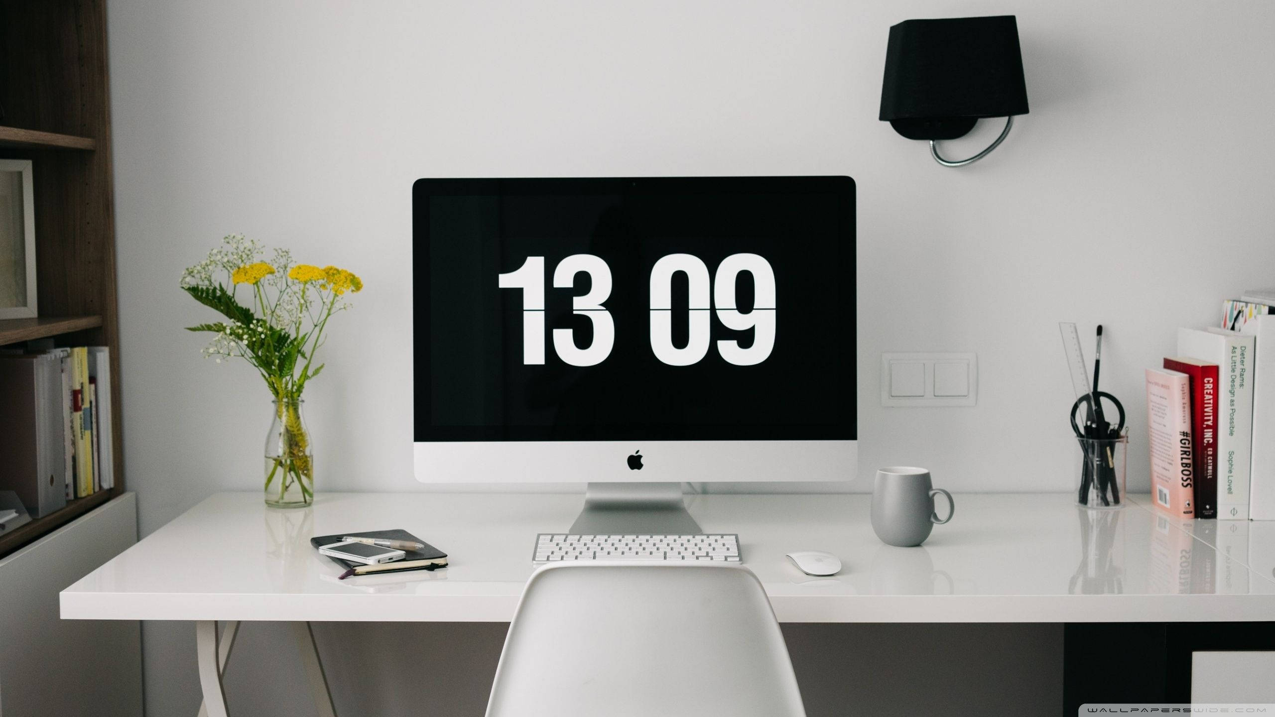 Working 桌布 壁紙 — 一個簡約的白色桌面設置，iMac 在黑色螢幕上顯示粗體白色數字，周圍環繞著書籍、鮮花和辦公用品。