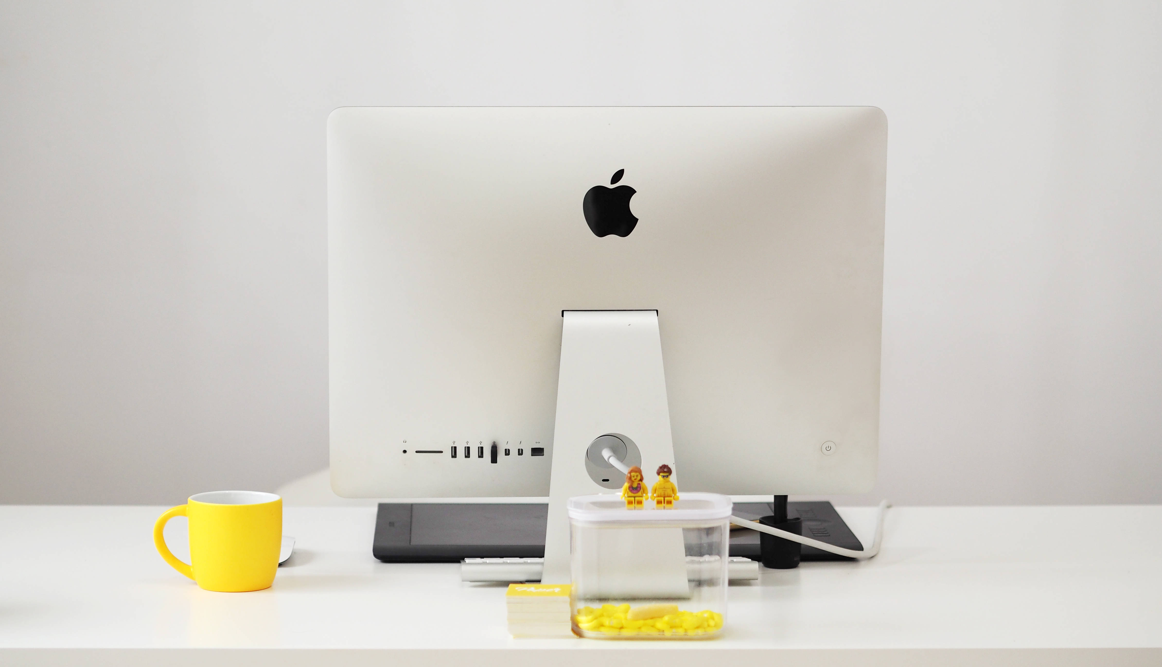 桌布 壁紙 — 一個簡約的白色桌面設置，配備 Apple iMac、一個亮黃色馬克杯，以及透明容器旁邊的細小 LEGO 人偶。