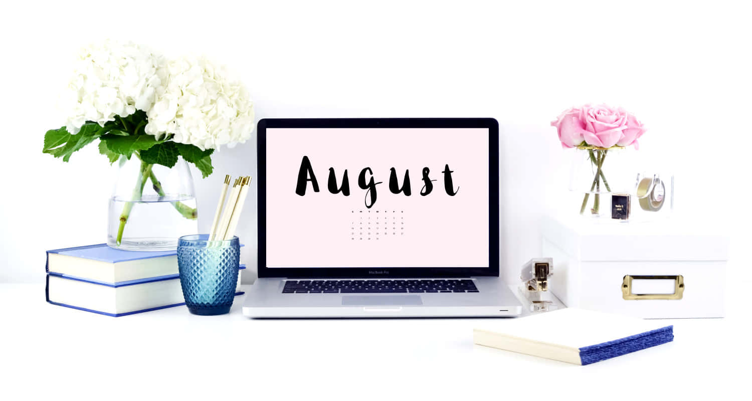 August 桌布 壁紙 — 一個簡約的白色桌面設置，配備一台顯示八月的筆記型電腦，搭配白色繡球花、粉紅玫瑰、書籍和文具。