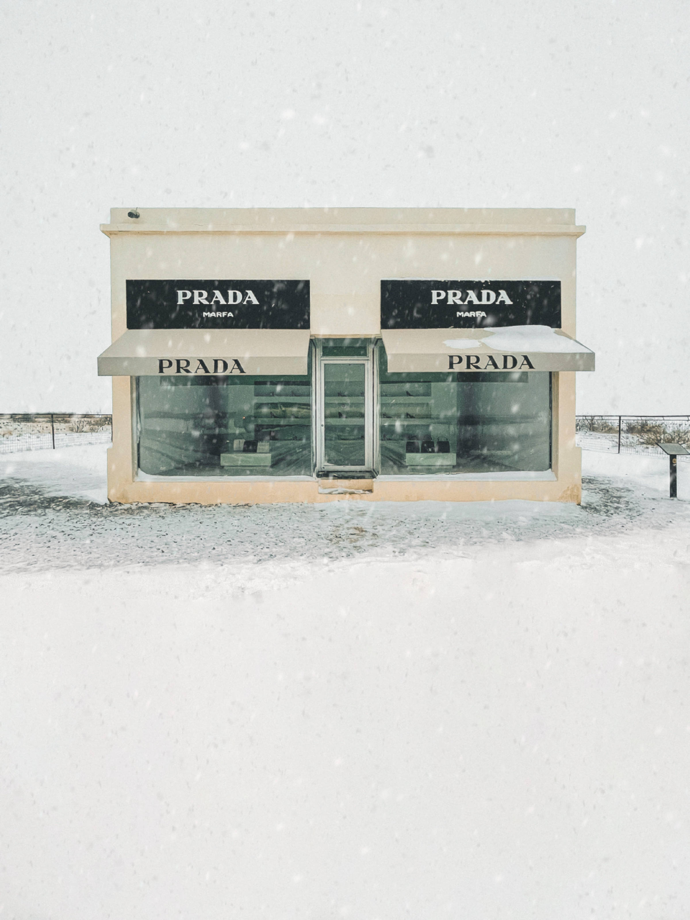Prada 桌布 壁紙 — 一個簡約的米色 Prada Marfa 藝術裝置獨自矗立在雪地沙漠景觀中，雪花飄落。
