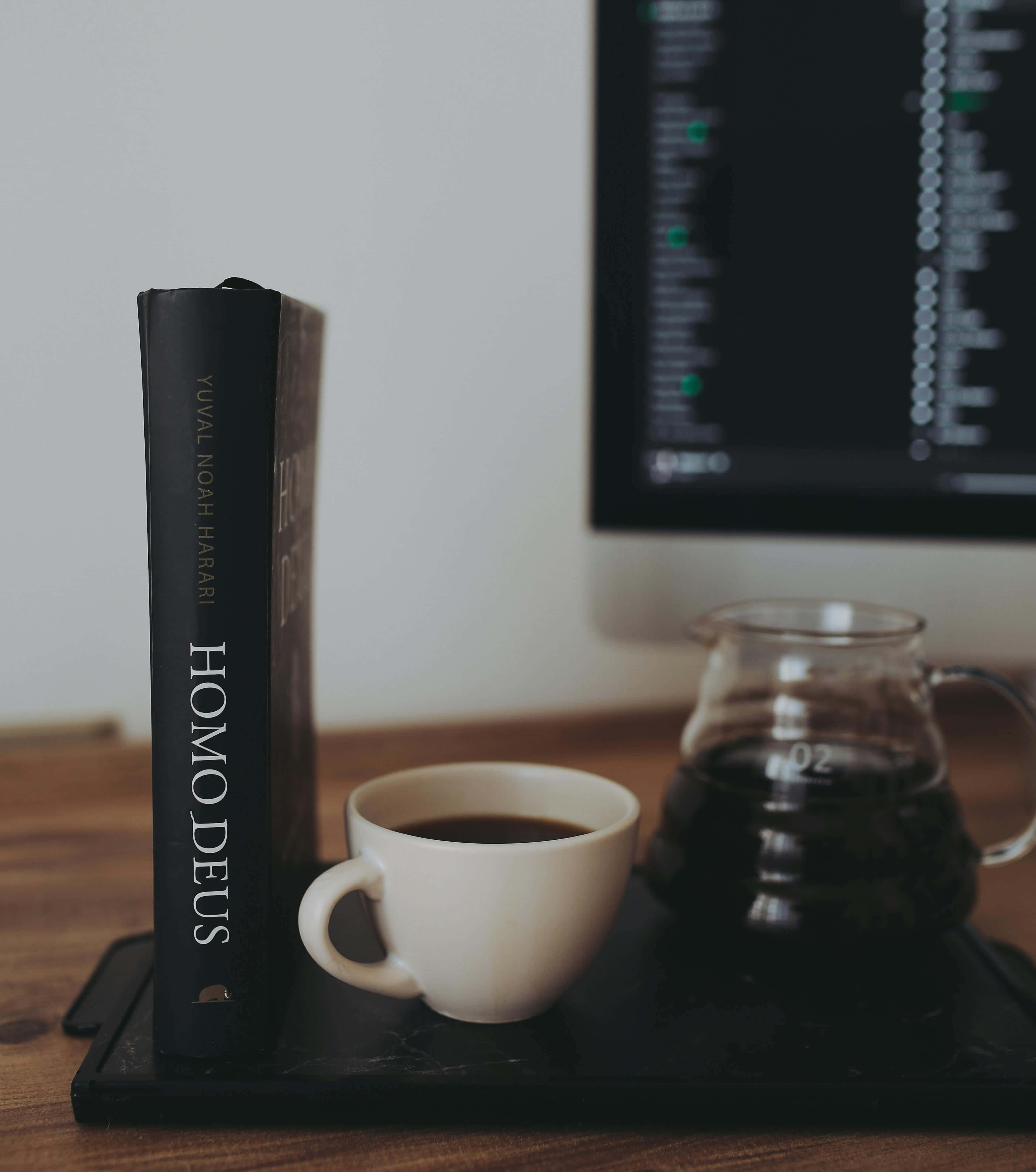 Dark Book Aesthetic 桌布 壁紙 — 一個簡約的辦公桌設置，旁邊有一本書、咖啡杯和手沖咖啡，電腦螢幕顯示程式碼。