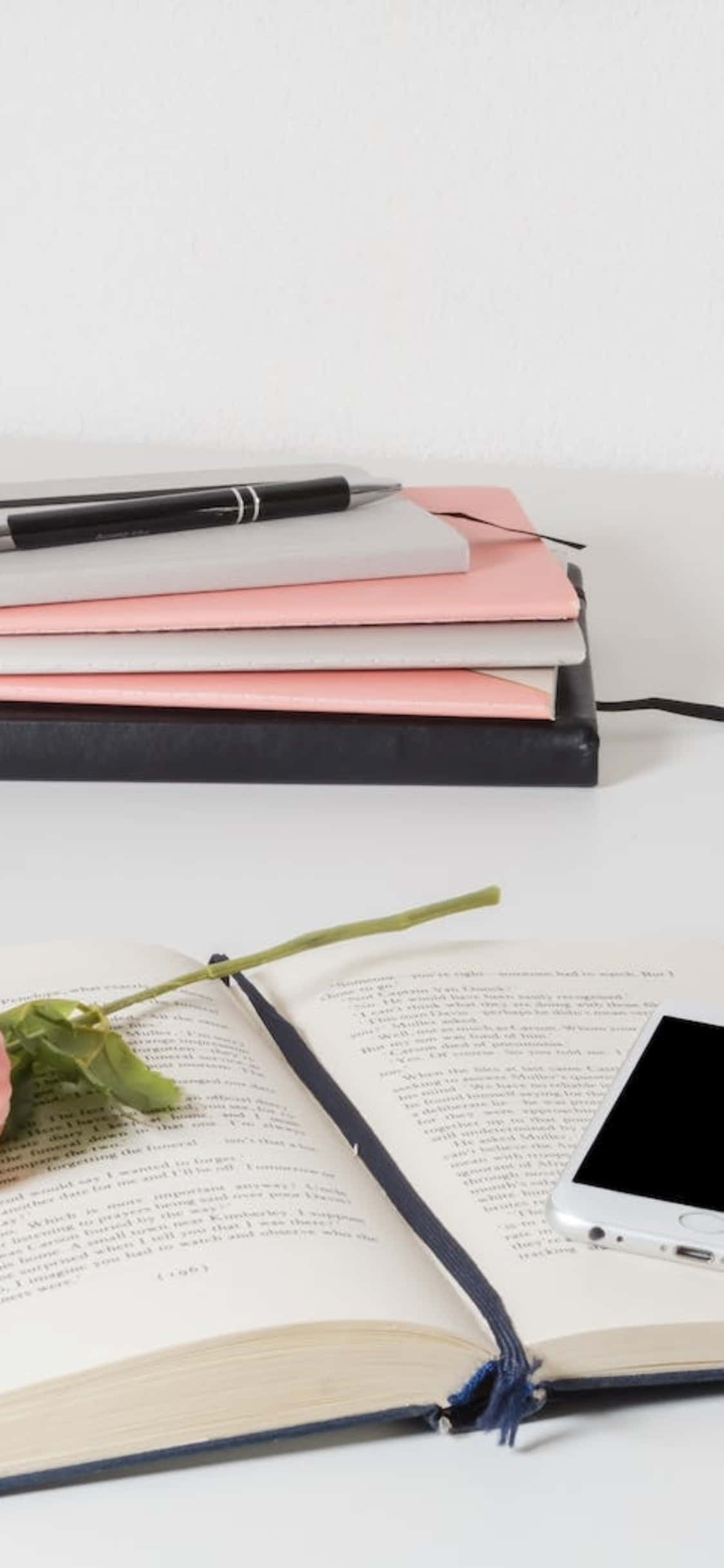 Desk 桌布 壁紙 — 一個簡約的辦公桌設置，有一本打開的書、一枝玫瑰莖、一枝筆和一部智慧型手機放在一堆筆記本上，背景是純白色的牆壁。