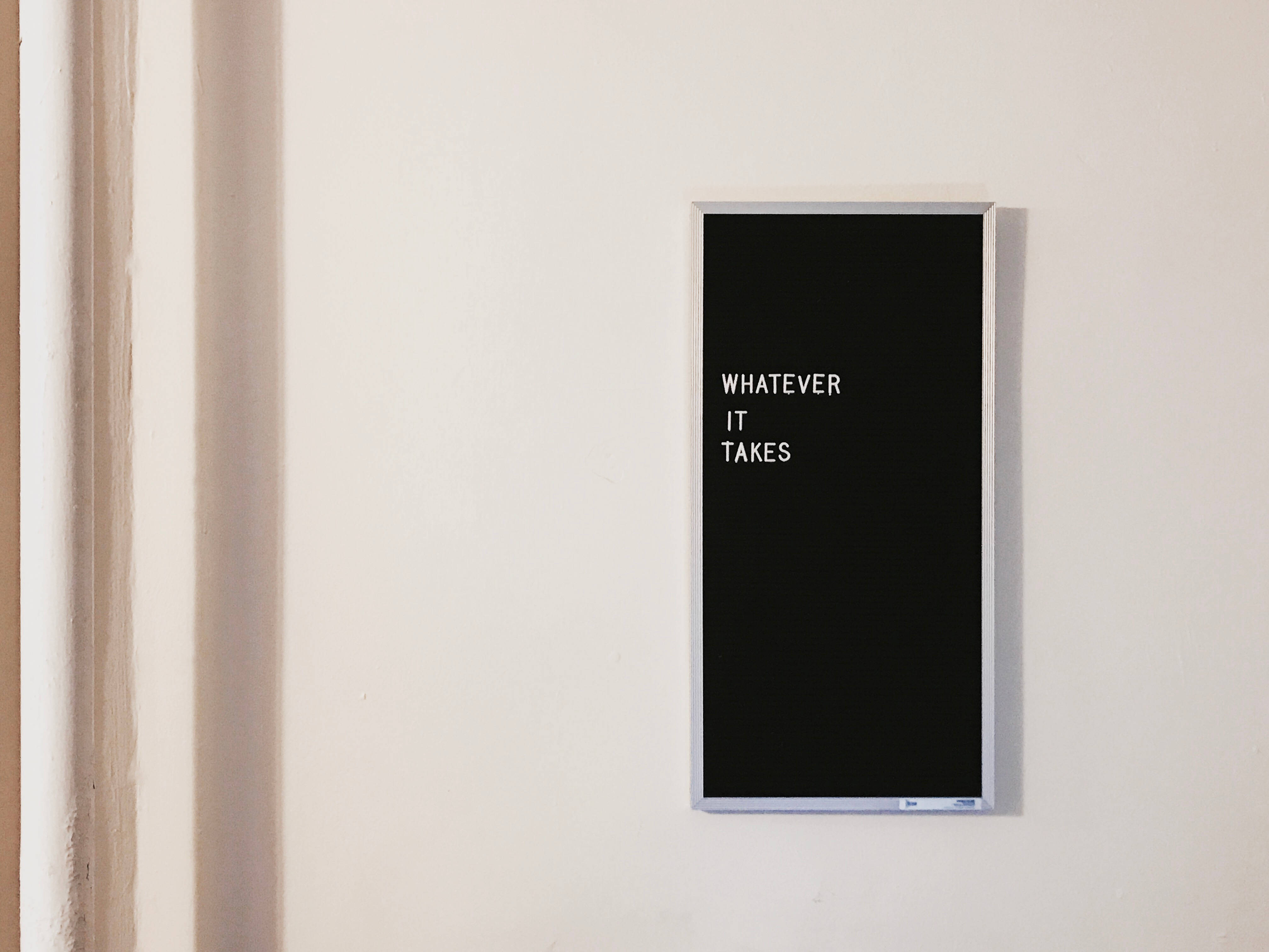 Brand 桌布 壁紙 — 一個簡約的黑色框標誌，白色文字寫著「WHATEVER IT TAKES」，安裝在純米白色牆上。