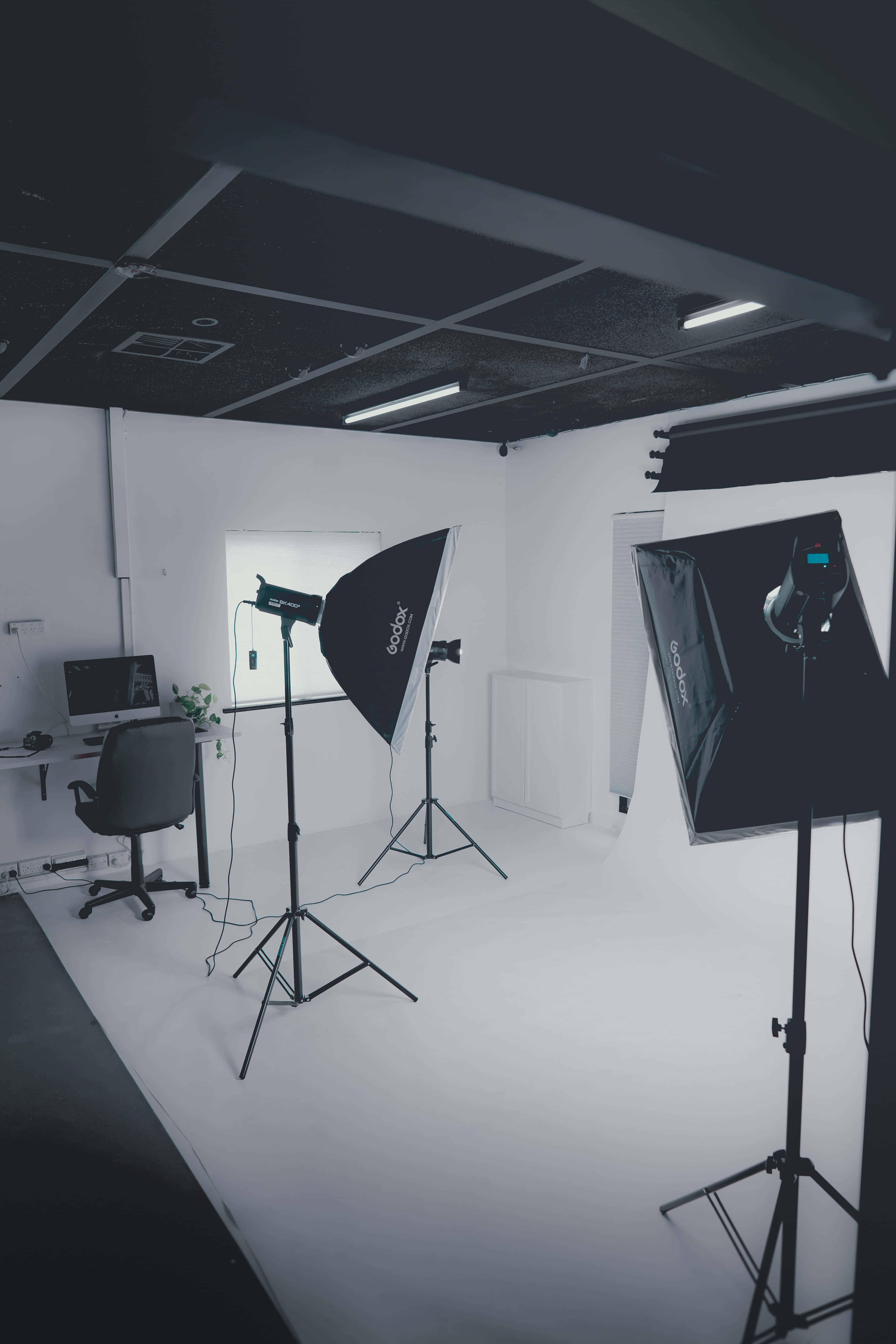 Photography Studio 桌布 壁紙 — 一個簡約風格攝影工作室，配有白色地板、黑色照明設備和螢光燈下的電腦桌。