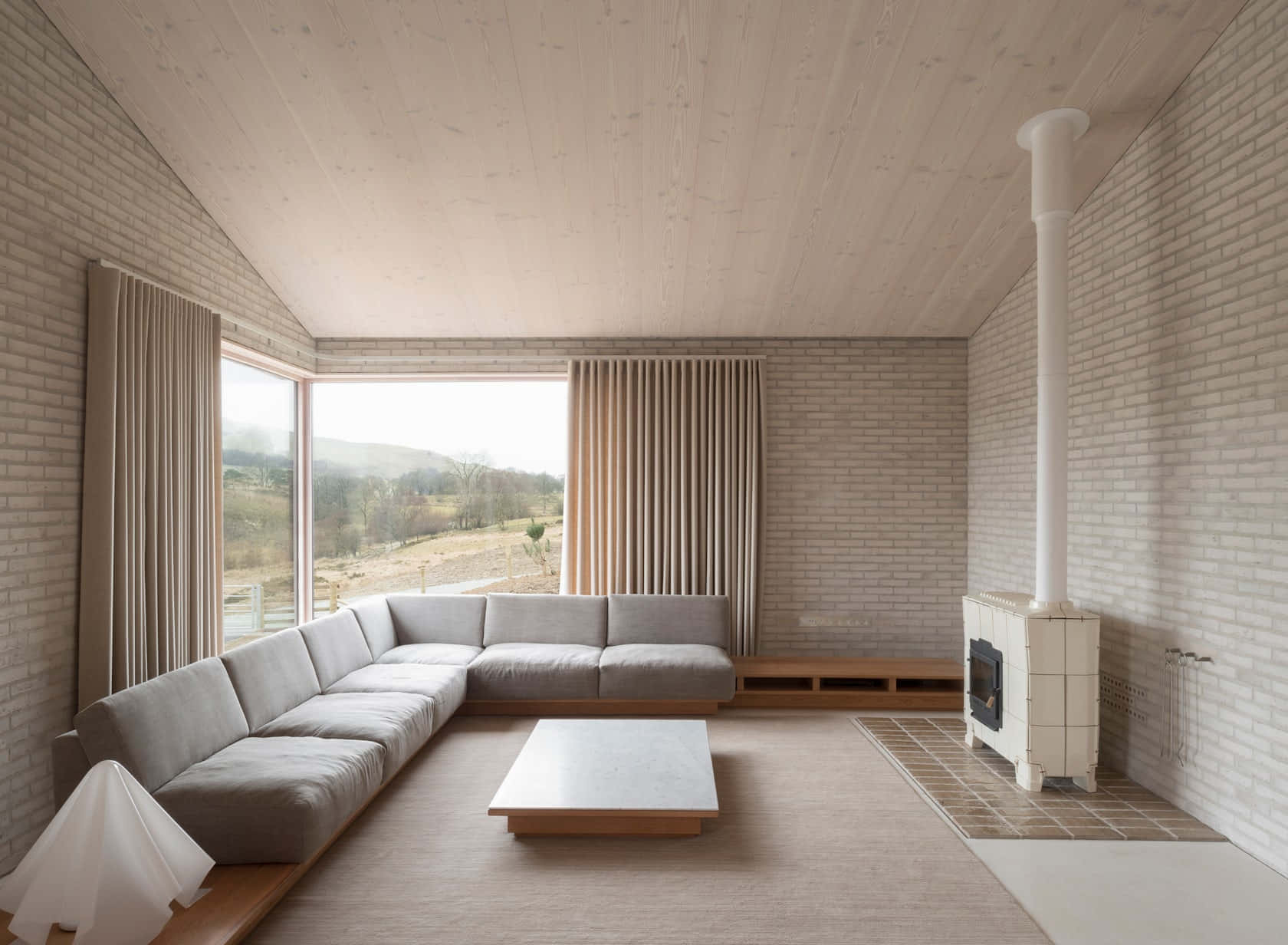Minimalist Architecture 桌布 壁紙 — 一個簡約風格的客廳，白色磚牆和木質天花板，配有一張大型組合沙發，面向窗外的連綿起伏山丘美景。
