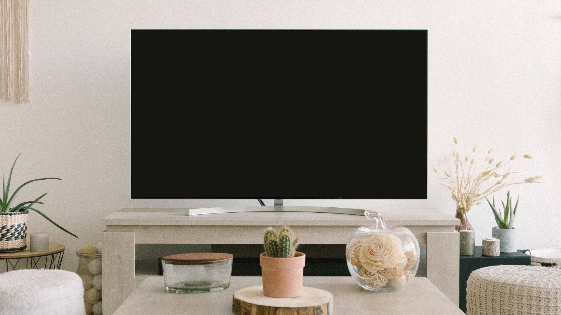 tv 桌布 壁紙 — 一個簡約風格的客廳，配備時尚電視機，置於淺色木架上，周圍環繞著盆栽仙人掌、乾花和舒適的針織腳凳。