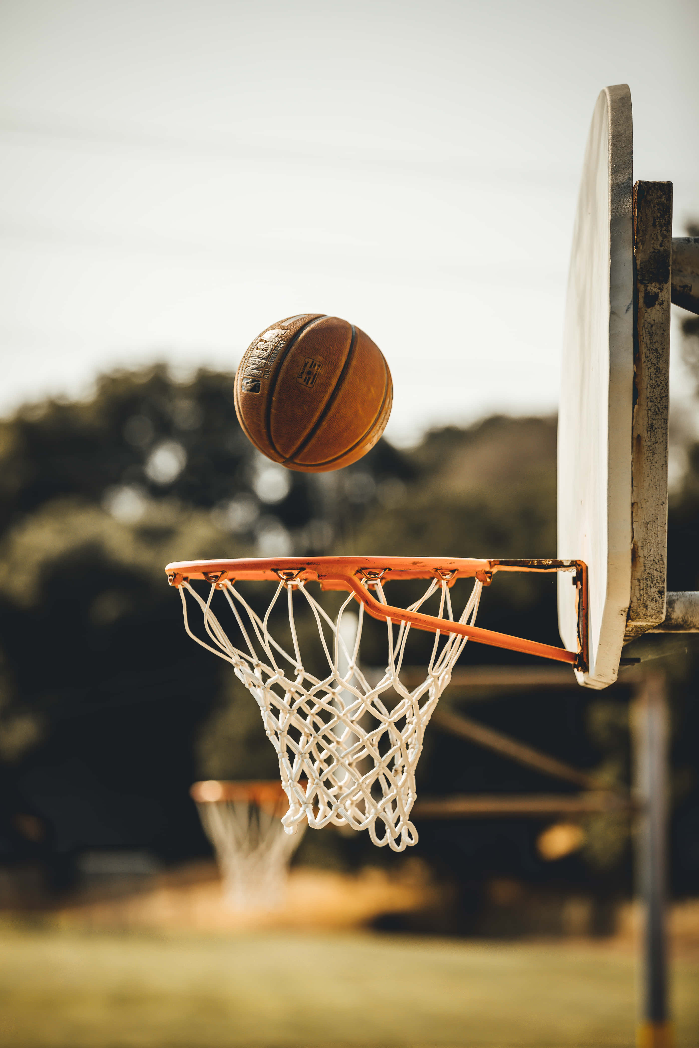 Best Sports 桌布 壁紙 — 一個籃球在半空中即將穿過一個磨損的籃框，帶有白色網，背景是模糊的戶外場景。
