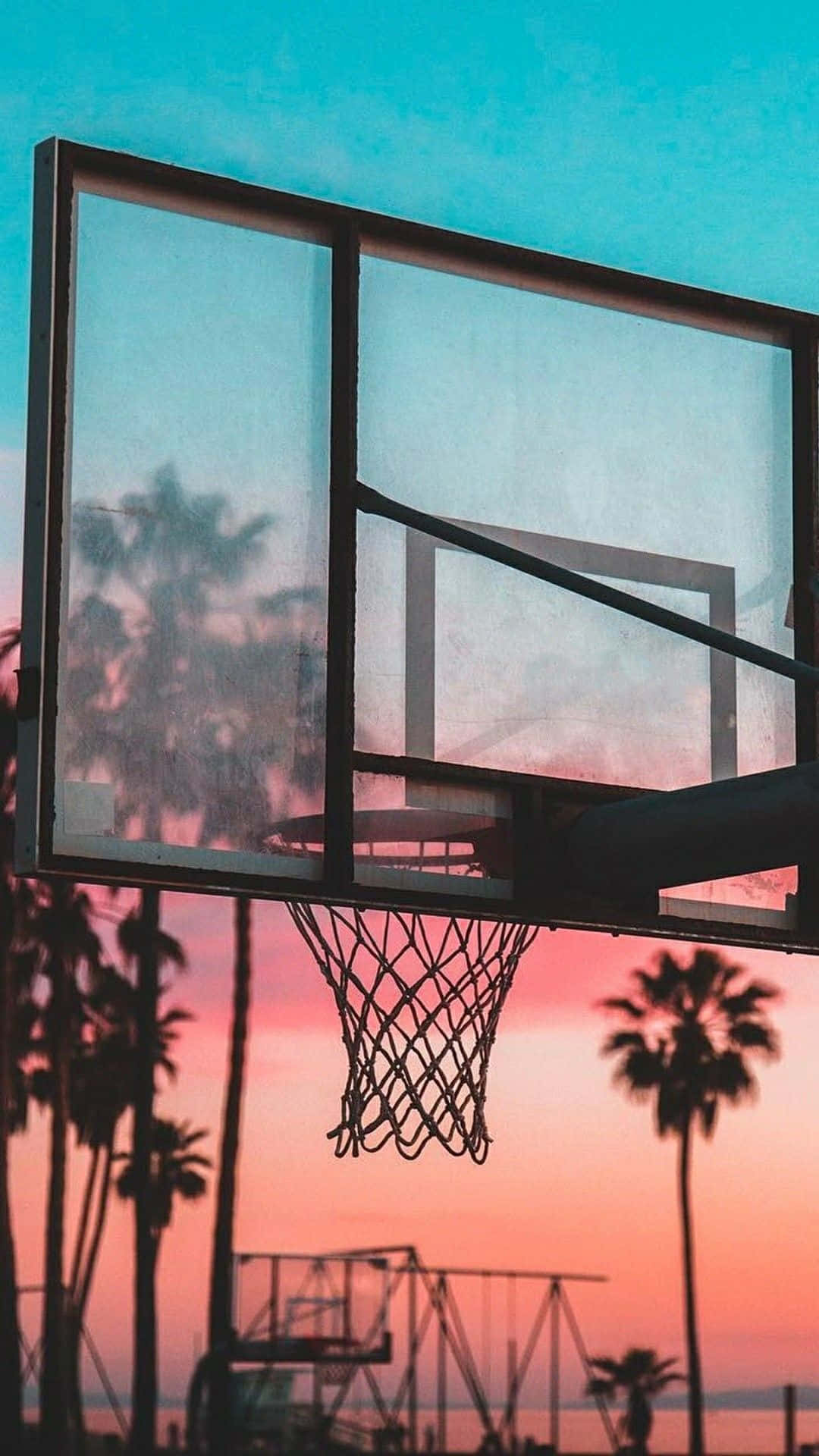 Basketball Aesthetic 桌布 壁紙 — 一個籃球框在充滿活力的日落天空下形成剪影，下方有棕櫚樹和遊樂場。