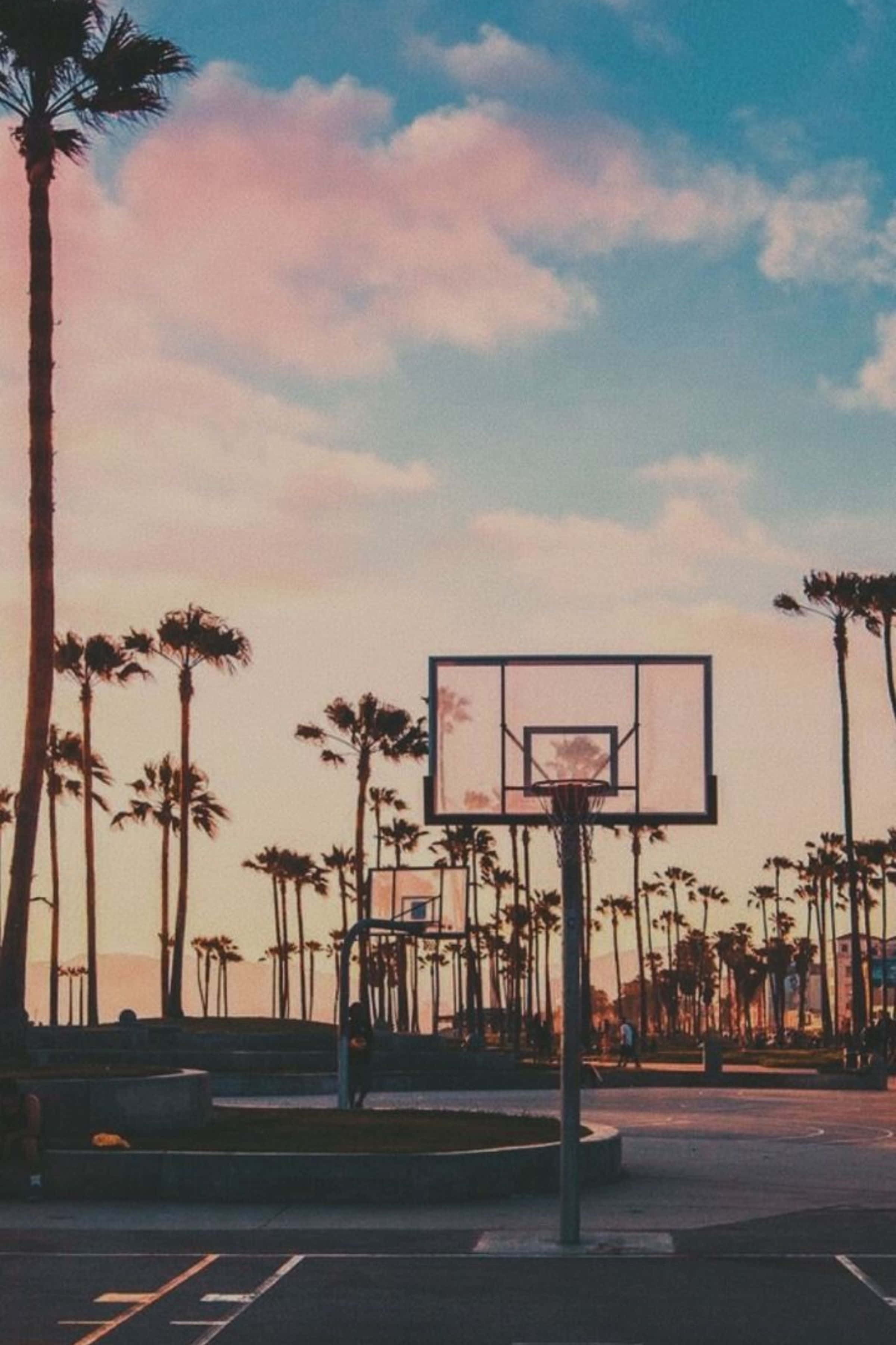 Life Aesthetic 桌布 壁紙 — 一個籃球框在柔和的雲彩和粉彩日落天空下，聳立在棕櫚樹之間。