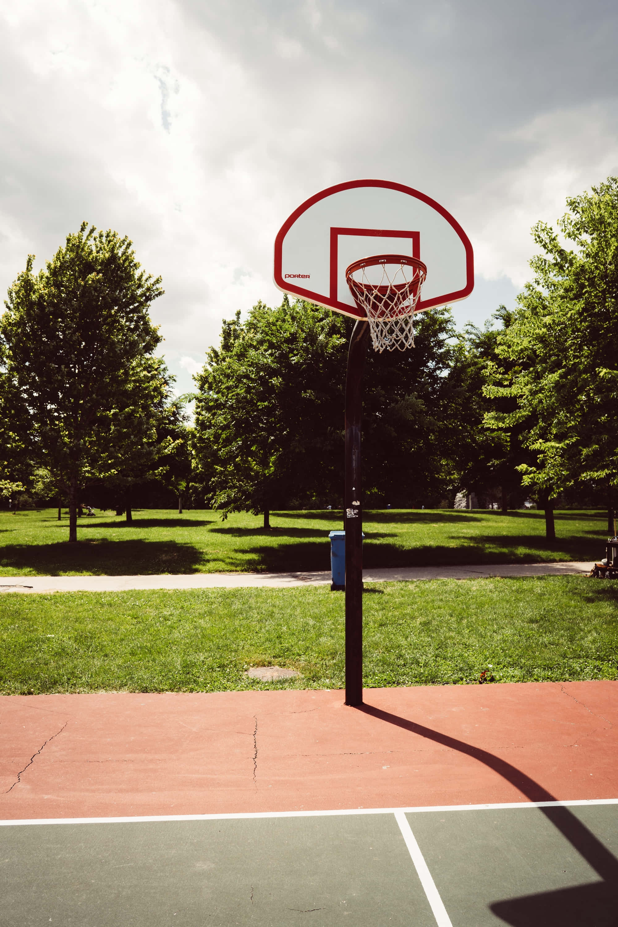 Basketball Court 桌布 壁紙 — 一個籃球框聳立在紅綠相間的球場上，周圍是茂密的綠樹，天空多雲。