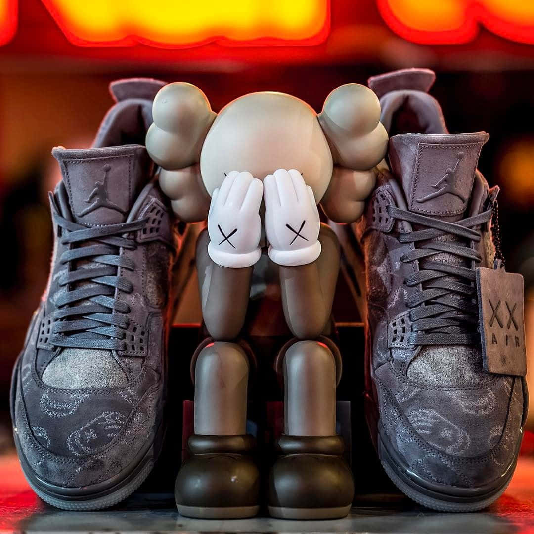 Kaws X Air Jordan 桌布 壁紙 — 一個米色KAWS公仔交叉雙手，坐在兩雙灰色Jordan運動鞋之間，下方是發光的紅色霓虹燈。