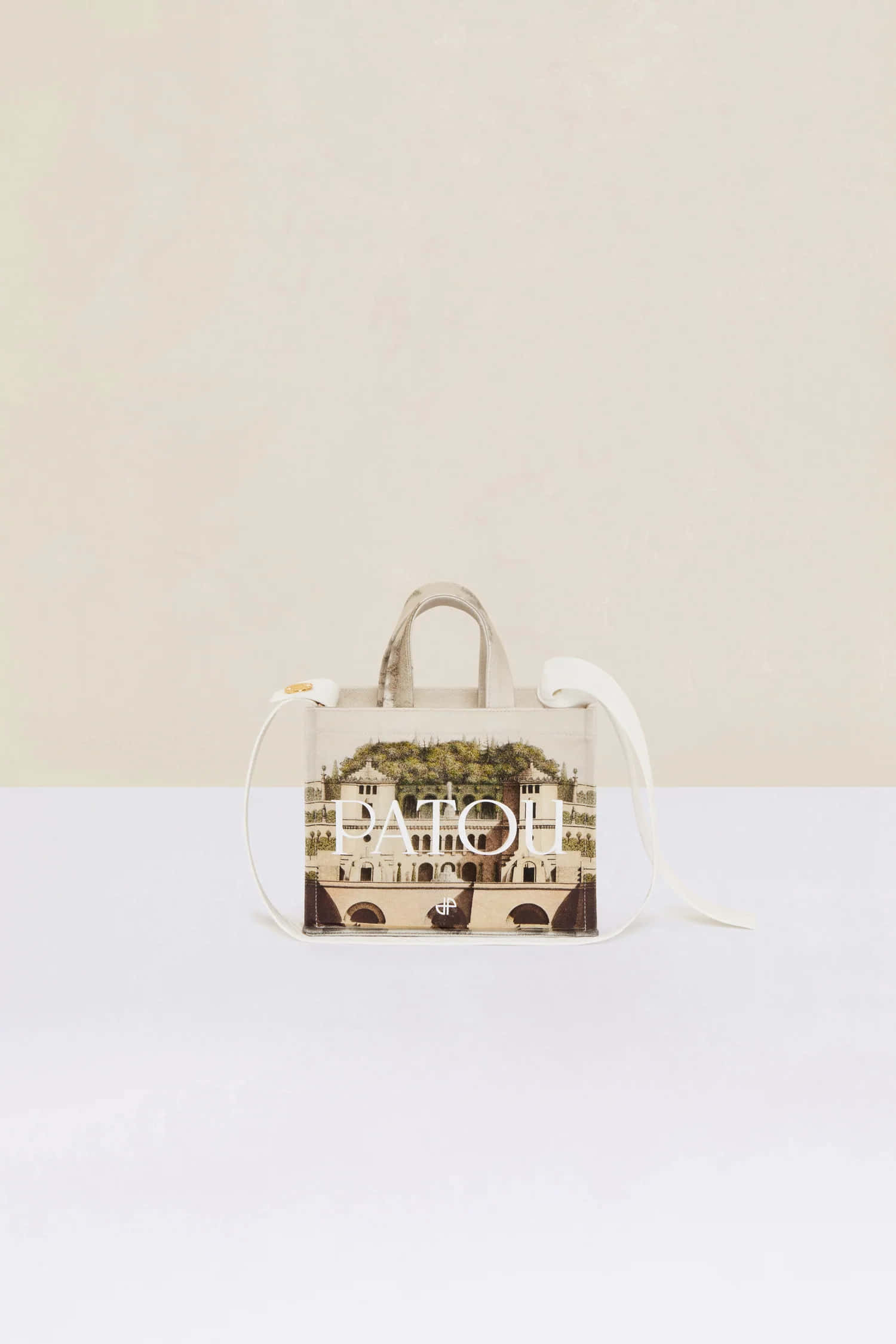 Handbag 桌布 壁紙 — 一個米色Patou手提包，印有復古建築場景，置於簡約白色表面上，背景為柔和奶油色。