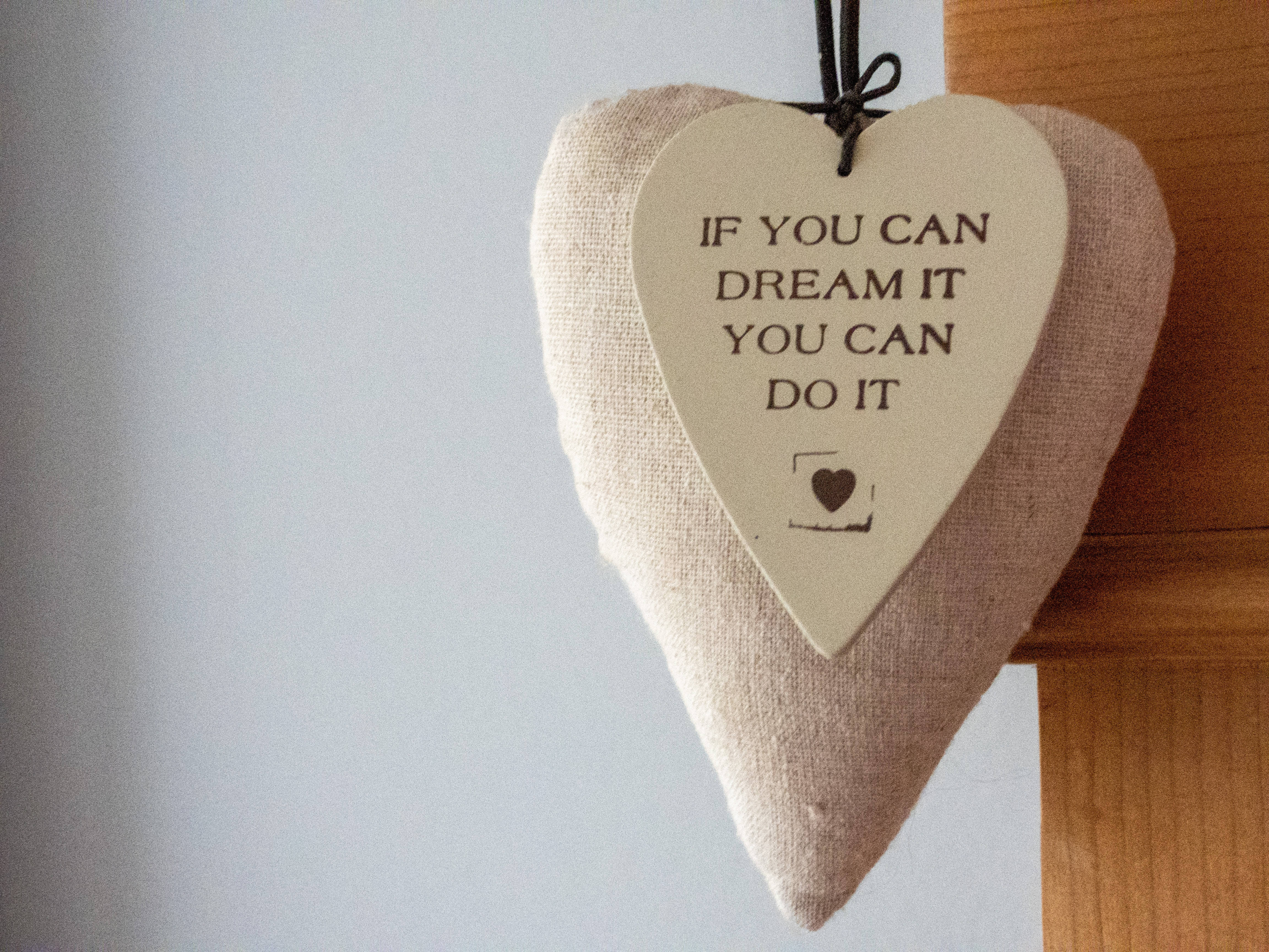 激勵 桌布 壁紙 — 一個米色亞麻心形裝飾品，上面寫著「if you can dream it you can do it」，掛在木樑上，背景是柔和的灰色牆壁。