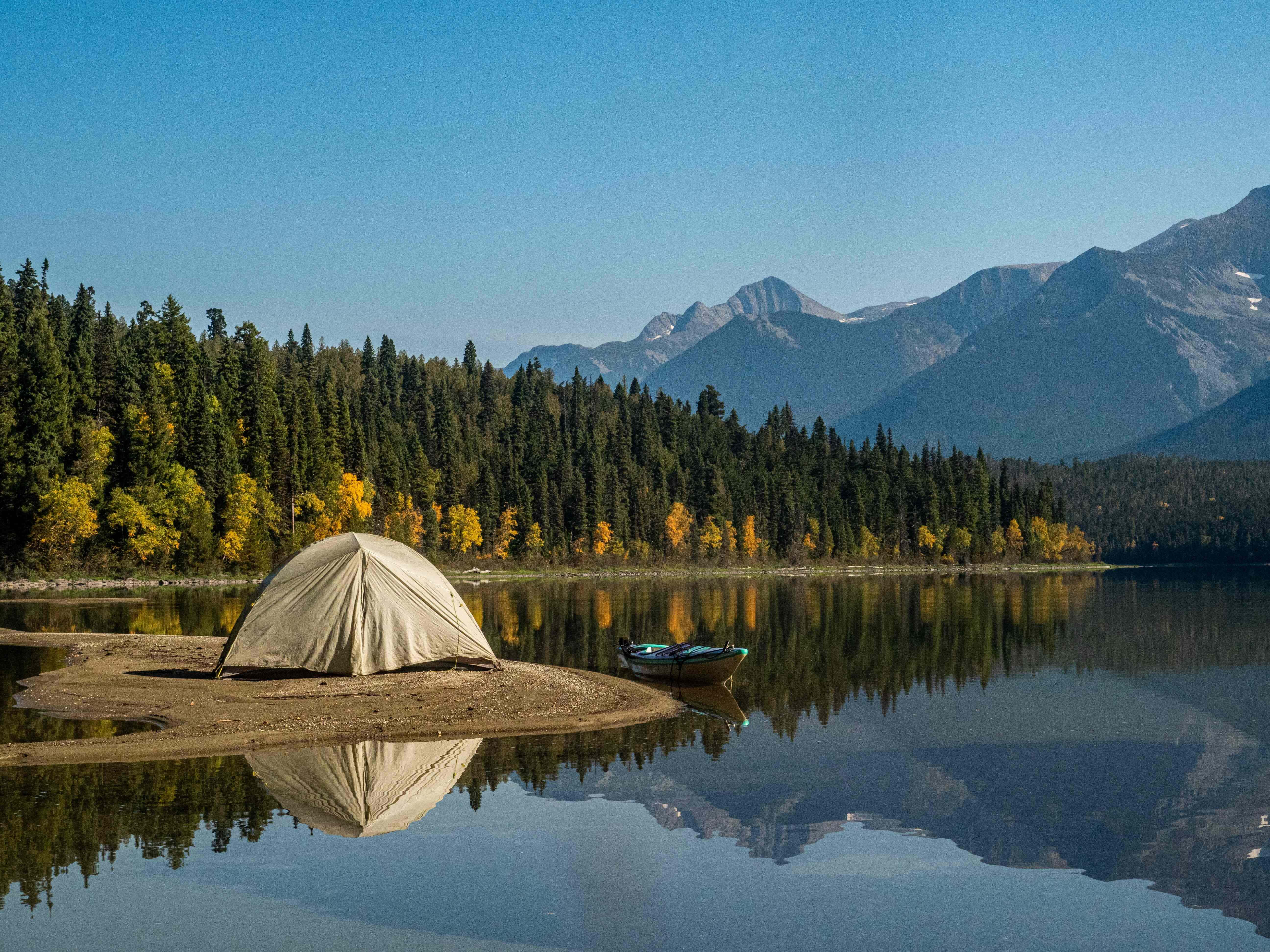 Camping 桌布 壁紙 — 一個米色帳篷位於平靜湖泊中的沙島上，附近有一艘小船，周圍是秋天的樹木和遠處白雪覆蓋的山脈，在清澈的藍天下。