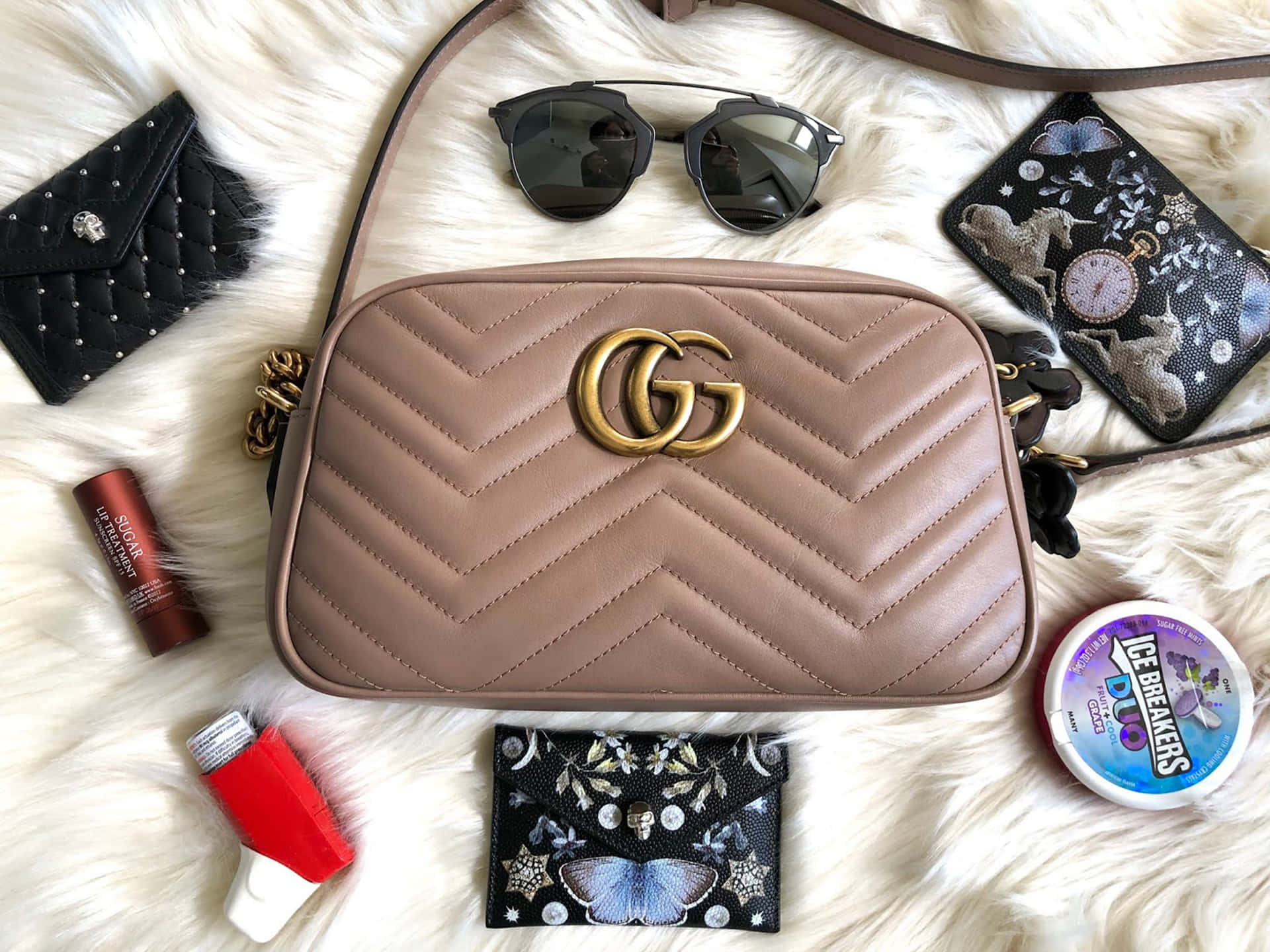 Gucci 桌布 壁紙 — 一個米色絎縫Gucci手袋放在蓬鬆的白色毛皮上，周圍環繞著太陽眼鏡、潤唇膏和裝飾性錢包。