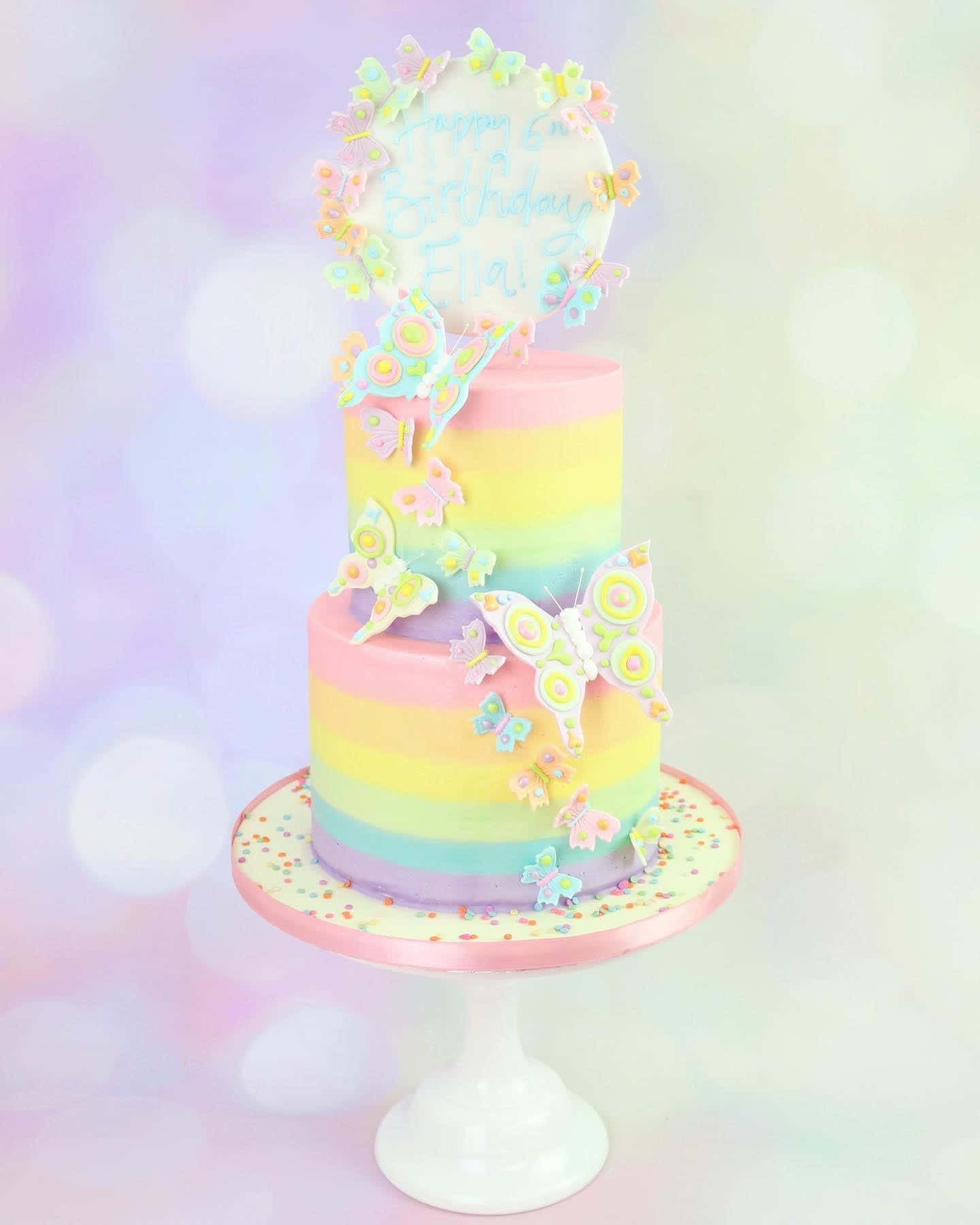 pastel 桌布 壁紙 — 一個粉彩彩虹分層生日蛋糕，裝飾著彩色蝴蝶和一個寫著「Happy Birthday Elial」的蛋糕插牌，放在白色架子上。