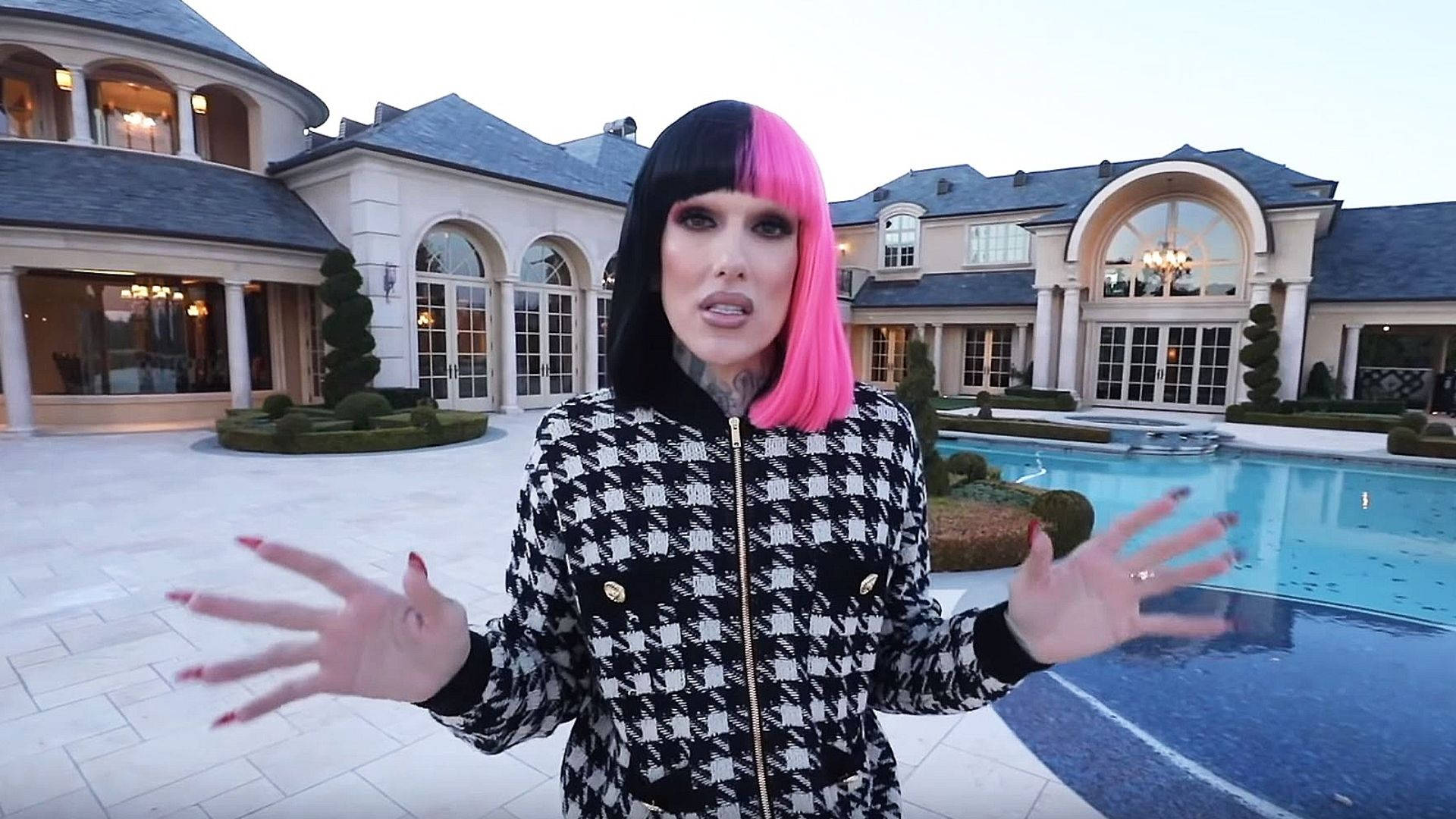 Jeffree Star 桌布 壁紙 — 一個粉紅和黑色頭髮的人穿著千鳥格外套，在黃昏時分於一個帶游泳池的豪華豪宅前做手勢。