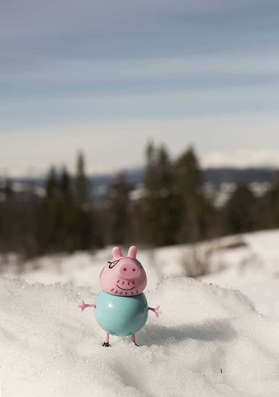 Daddy Pig 桌布 壁紙 — 一個粉紅色Peppa Pig玩具站在新雪中，背景有模糊的常綠樹和遠山，天空是淡藍色。