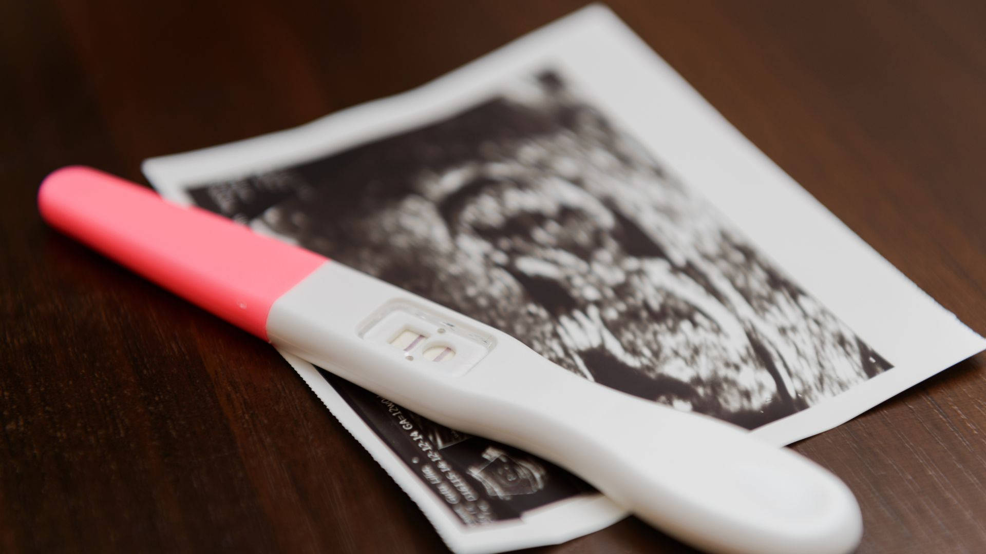 Pregnant 桌布 壁紙 — 一個粉紅色和白色的陽性懷孕測試棒放在一張印刷的超音波圖像上，置於深色木桌上。