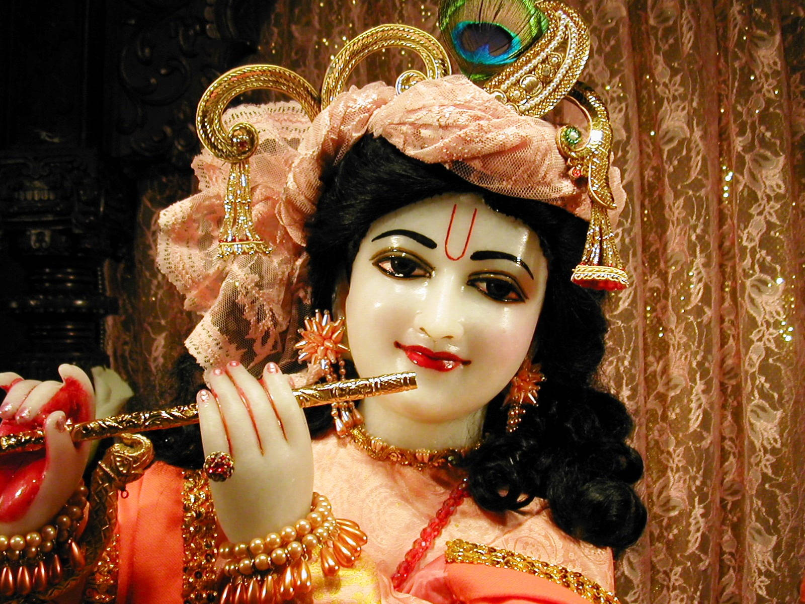 krishna god 桌布 壁紙 — 一個粉紅色和金色裝飾的克里希納神像，戴著孔雀羽毛皇冠，在閃閃發光的窗簾背景下吹奏長笛。