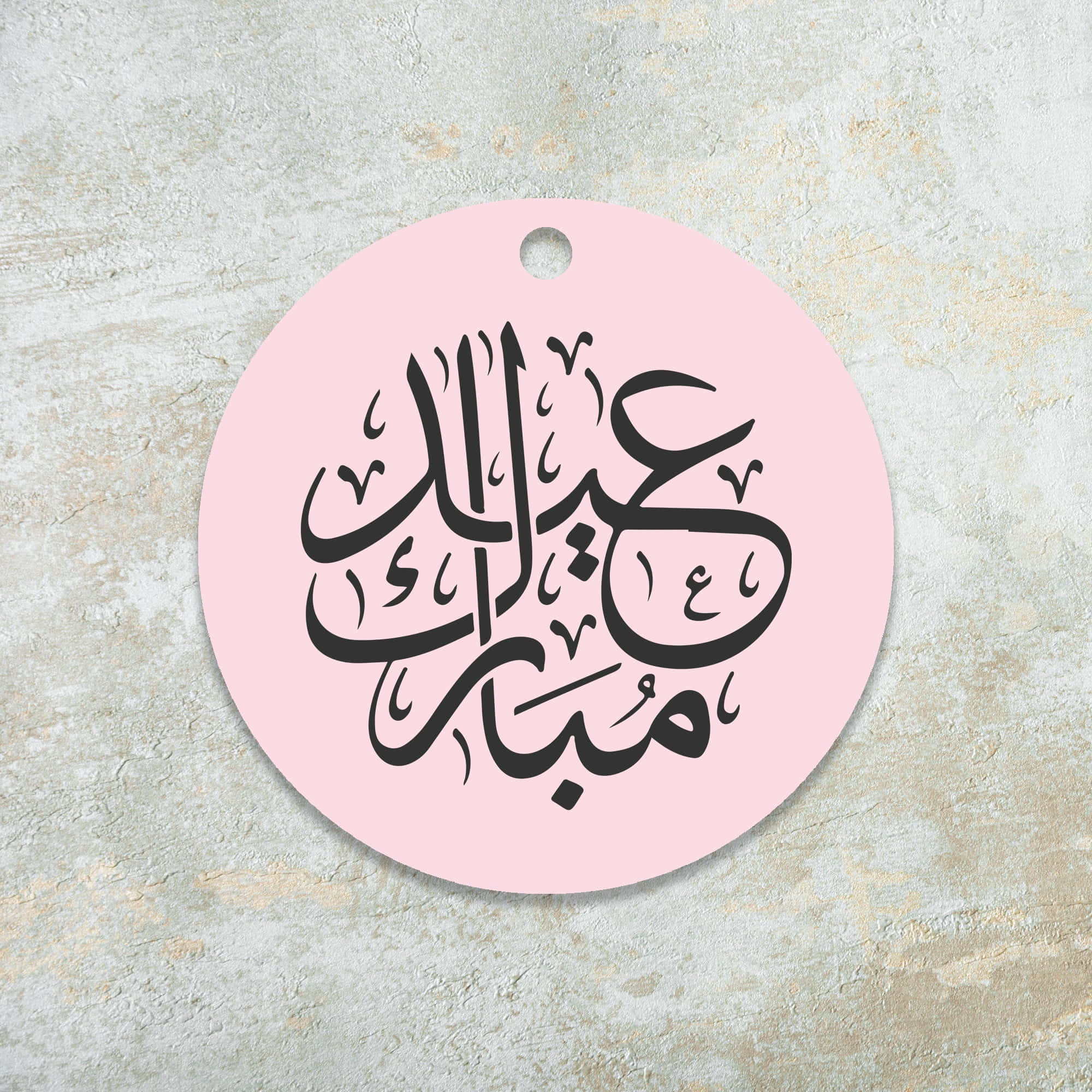 Eid Mubarak 桌布 壁紙 — 一個粉紅色圓形標籤，帶有優雅的黑色阿拉伯書法，掛在紋理米色牆上。