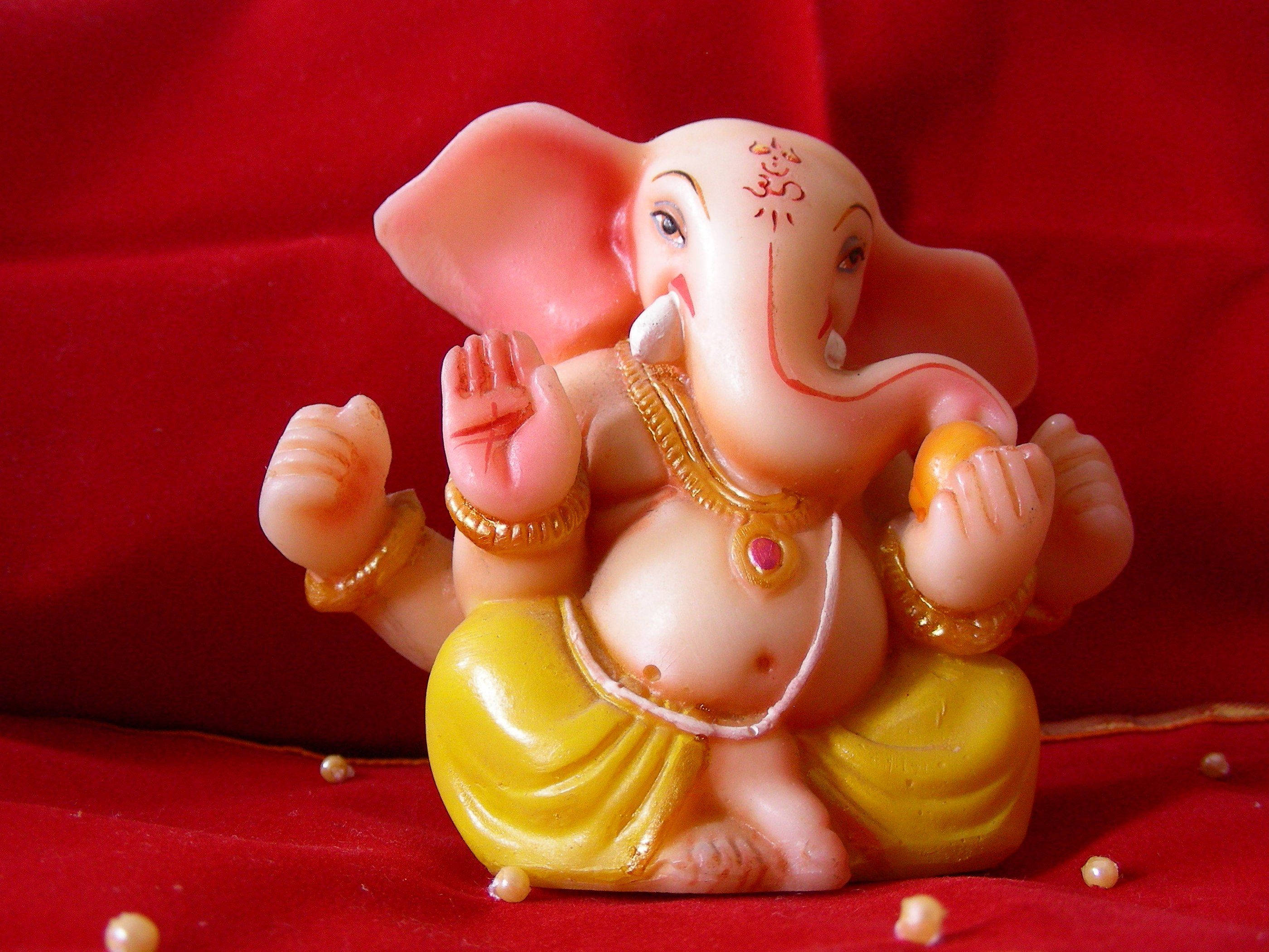 Ganesh 桌布 壁紙 — 一個粉紅色塑膠象神雕像，佩戴金色珠寶和黃色腰布，坐在華麗的紅色天鵝絨表面上，周圍散落著珍珠。