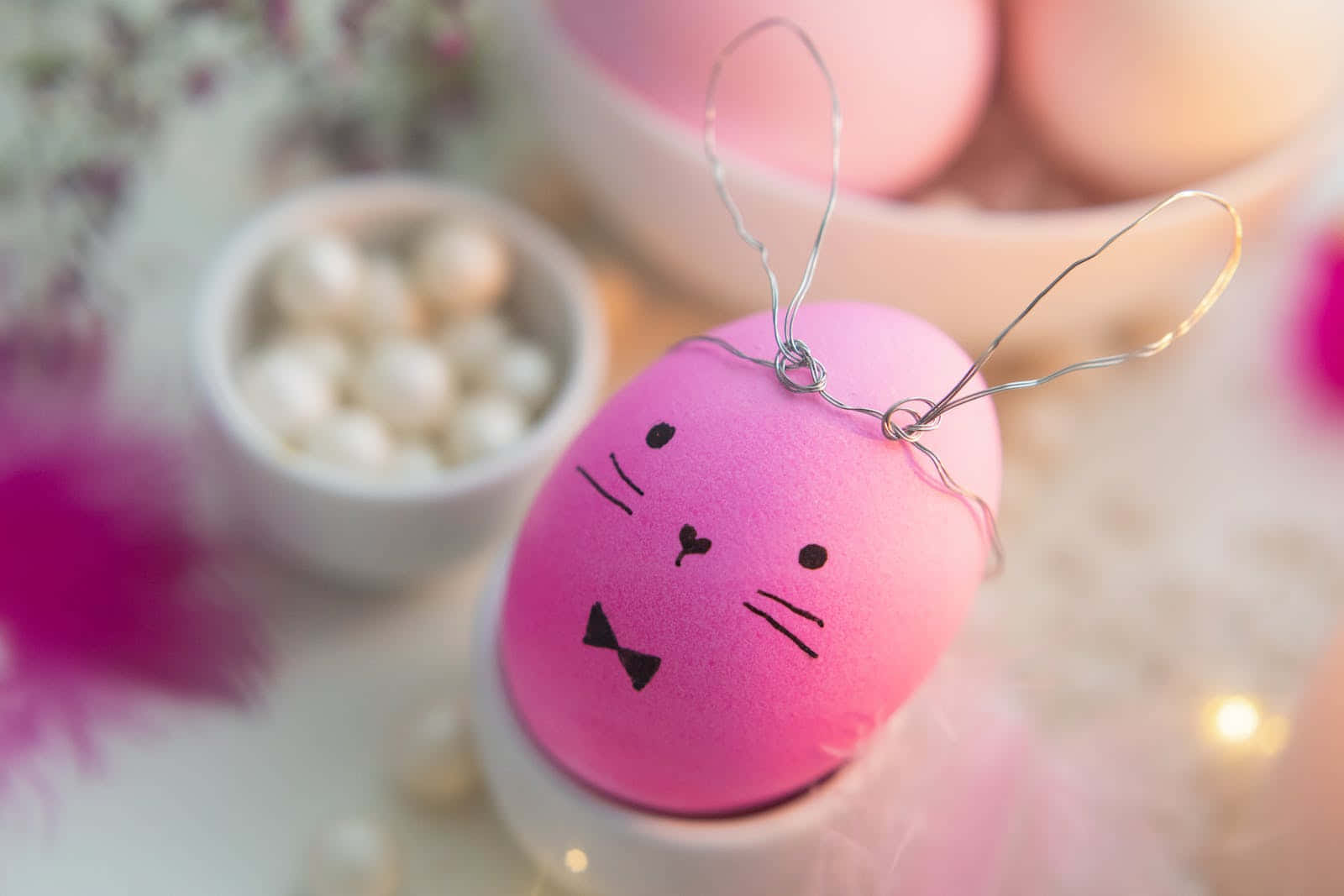 Pastel Easter 桌布 壁紙 — 一個粉紅色復活節蛋，帶有鐵絲兔耳和可愛貓臉，放在白色蛋杯中，周圍環繞著珍珠和柔和粉紅色裝飾。