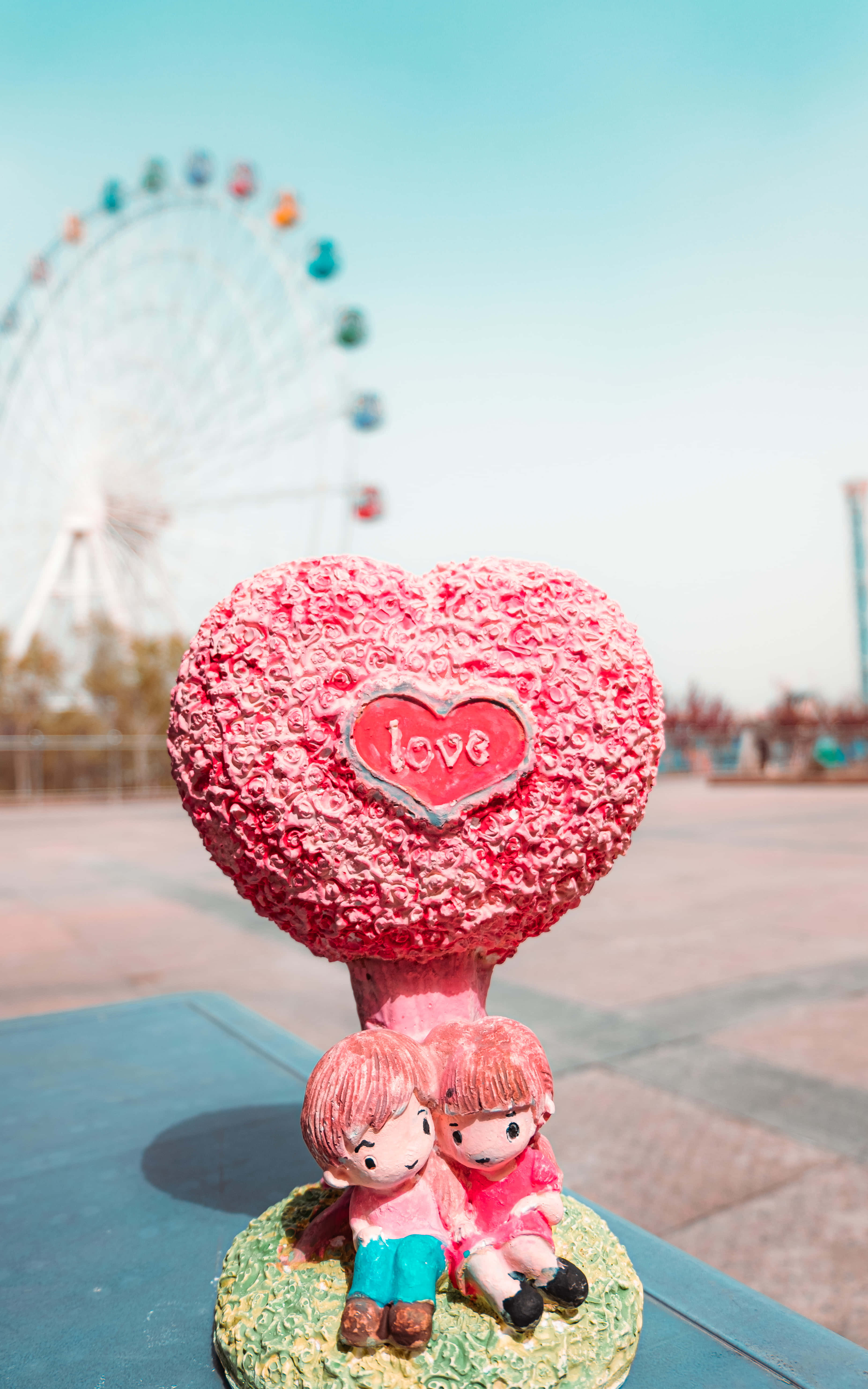 可愛的愛情 桌布 壁紙 — 一個粉紅色心形雕塑，上面刻有「love」，放在兩個卡通人物上面，背景是晴朗的藍天和摩天輪。