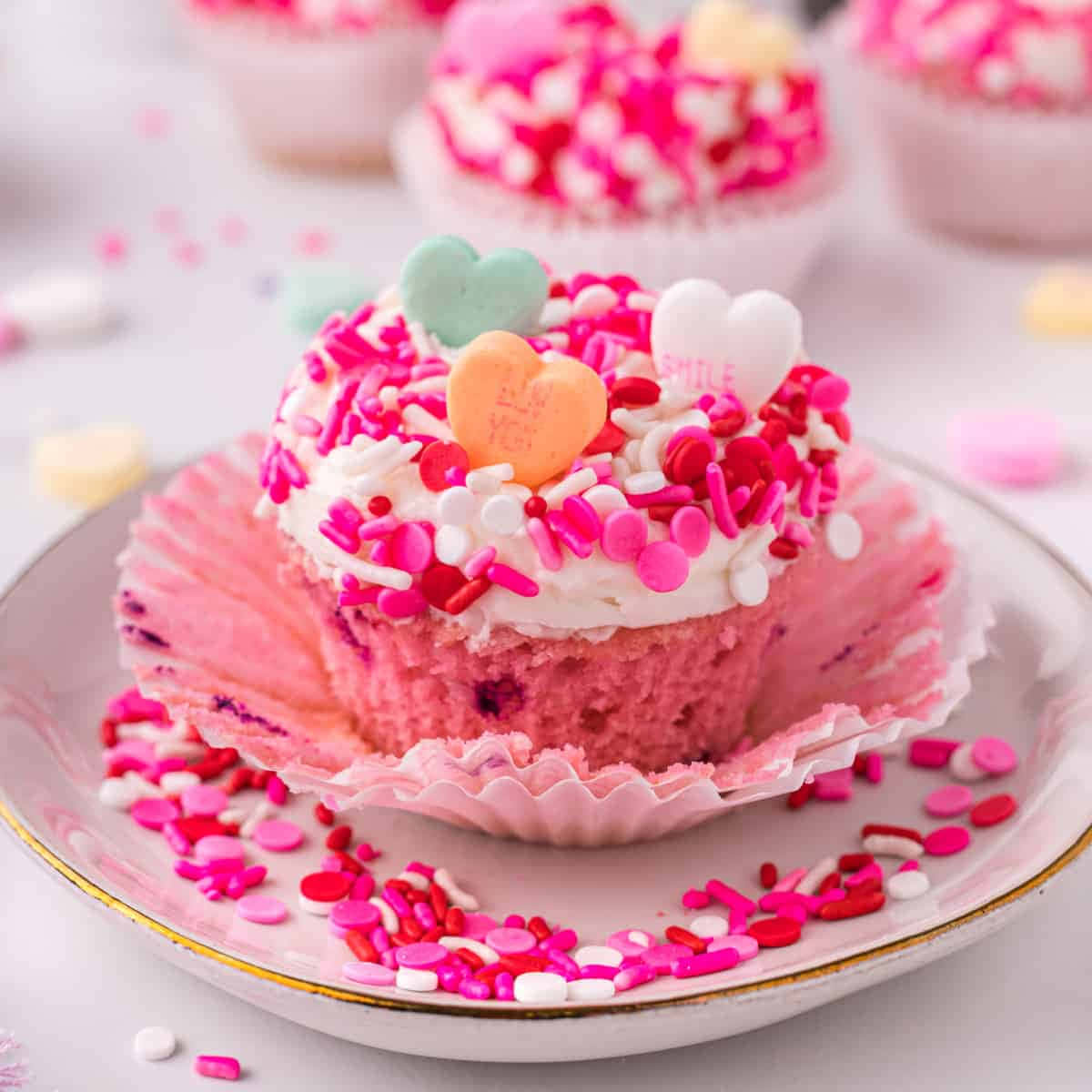 Pink Cupcakes 桌布 壁紙 — 一個粉紅色杯子蛋糕的特寫，上面有白色糖霜和色彩繽紛的心形糖粒，放在裝飾盤上。