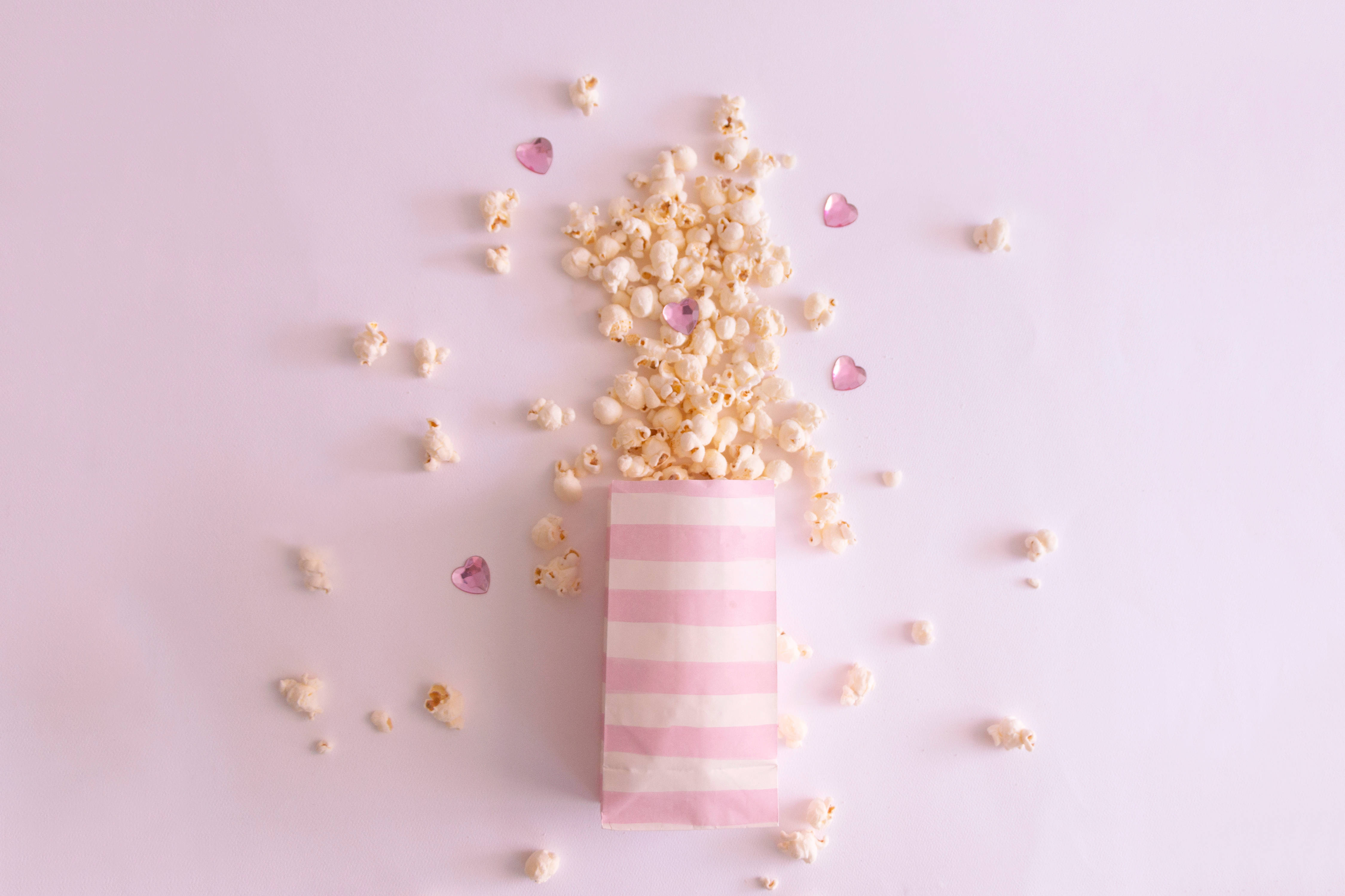 girly pink aesthetic 桌布 壁紙 — 一個粉紅色條紋袋，裝滿爆米花和散落的心形糖粒，置於柔和的粉彩背景上。