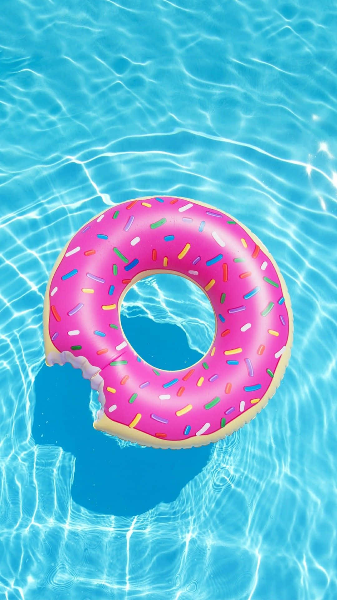 Trendy Summer iPhone 桌布 壁紙 — 一個粉紅色灑糖甜甜圈造型浮床，上面有咬痕，漂浮在清澈藍色泳池水中，帶有漣漪反射。