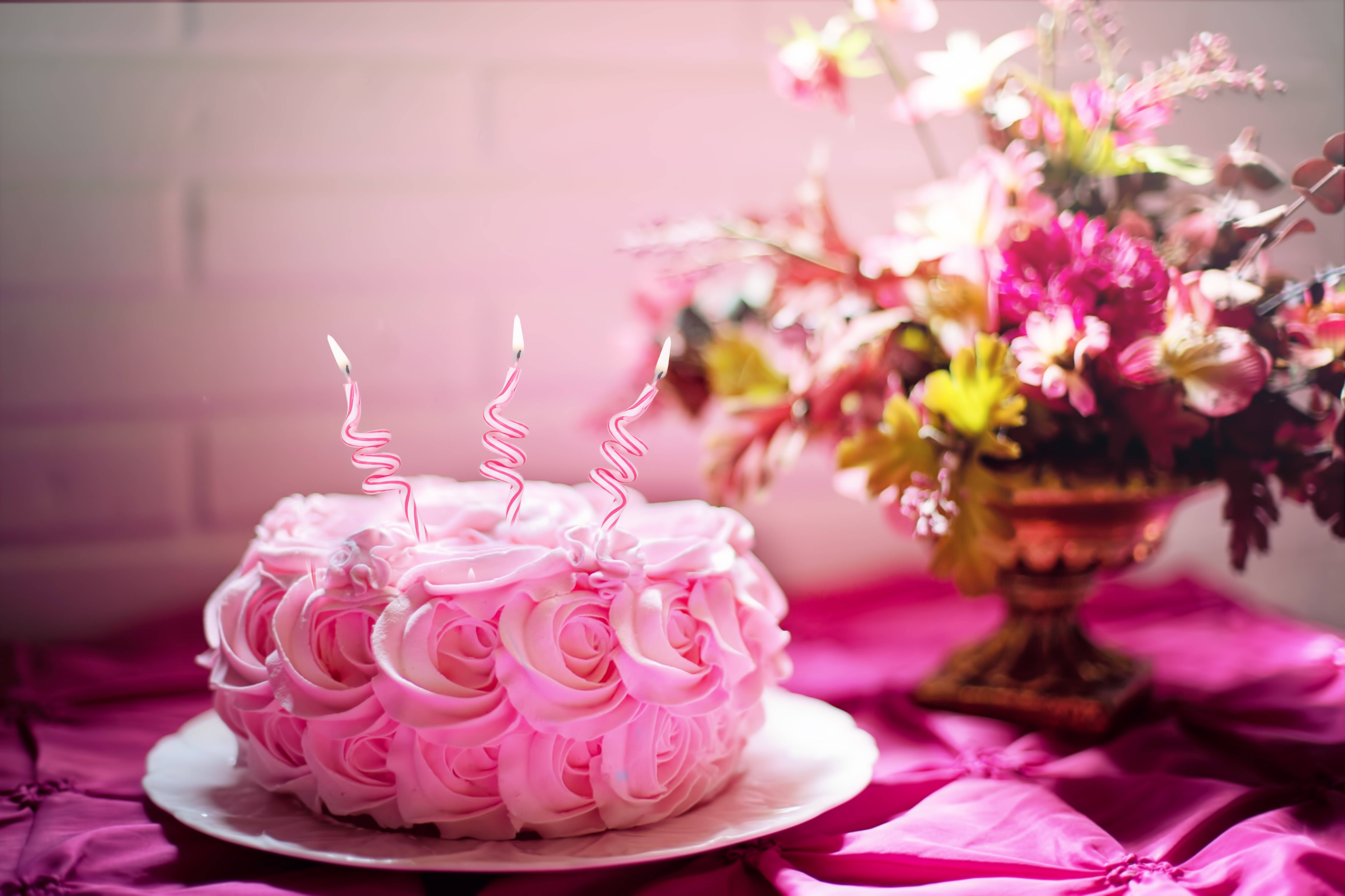 Birthday Cake 桌布 壁紙 — 一個粉紅色玫瑰圖案生日蛋糕，上面插著三根點燃的螺旋蠟燭，旁邊放著一個鮮豔的花藝佈置，置於洋紅色緞布上。
