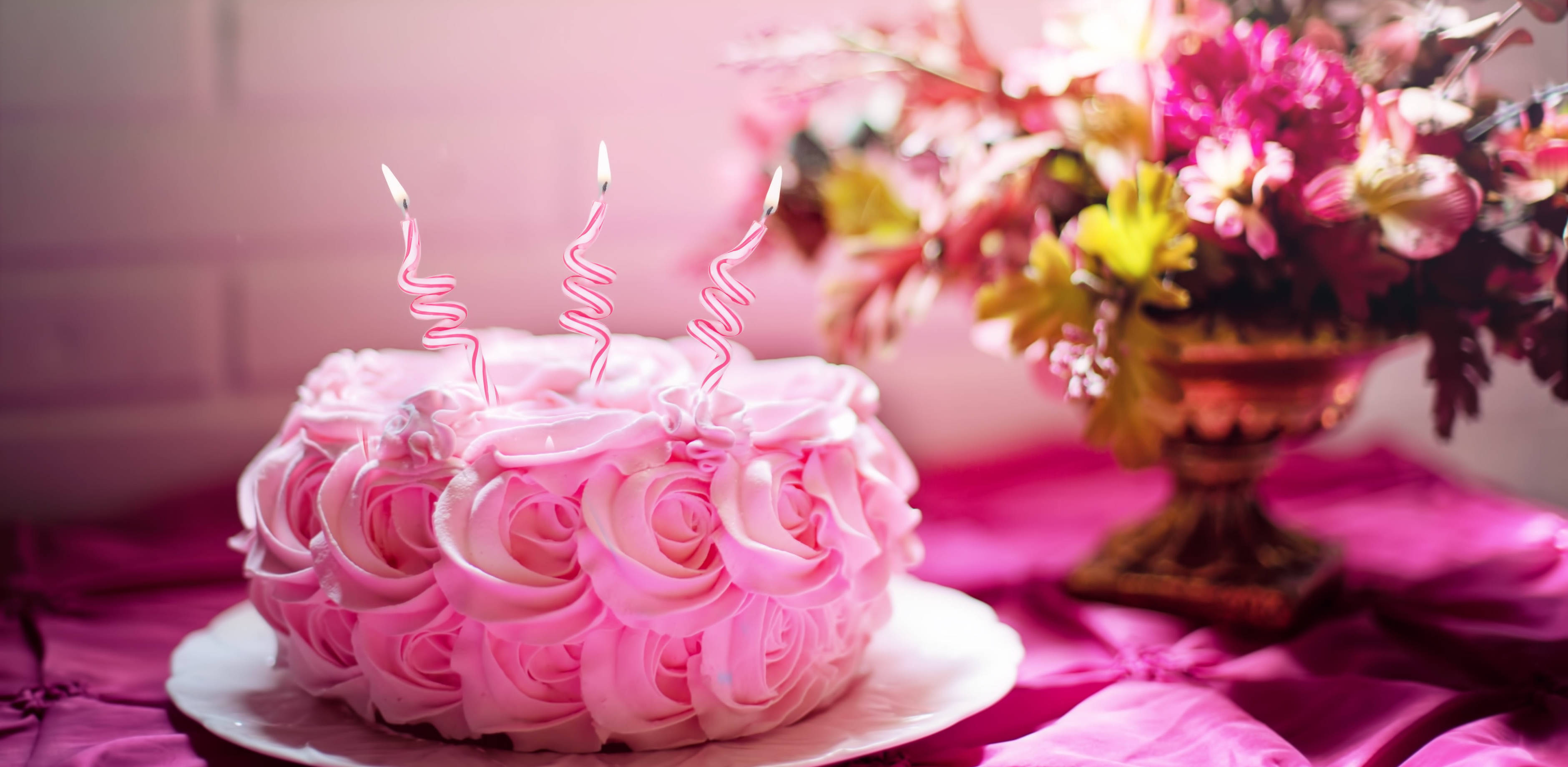 Happy Birthday Flowers 桌布 壁紙 — 一個粉紅色玫瑰形狀的蛋糕，上面插著三根點燃的蠟燭，放在洋紅色桌布上，旁邊有花卉佈置。