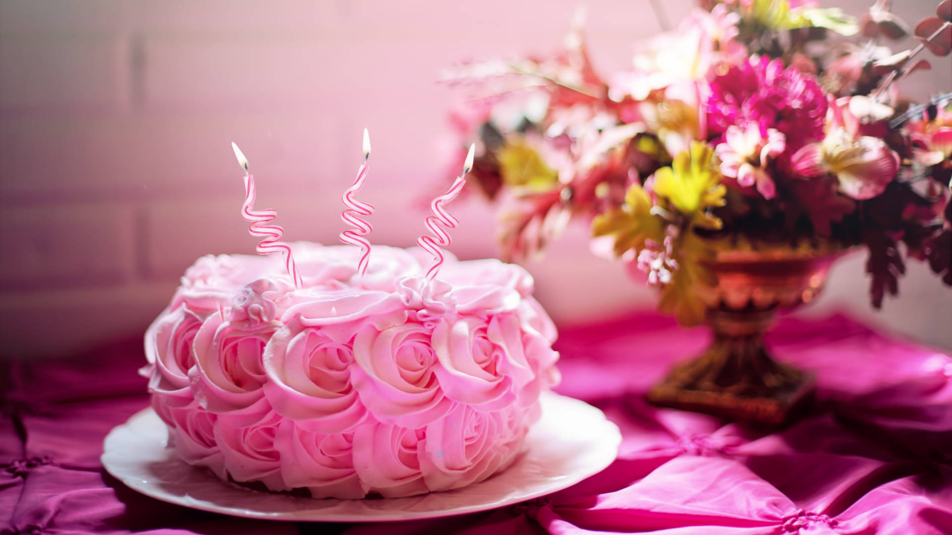Happy Birthday Flower 桌布 壁紙 — 一個粉紅色玫瑰形狀的蛋糕，上面有三根點燃的螺旋蠟燭，放在一個洋紅色的桌布上，旁邊有一個花卉佈置。