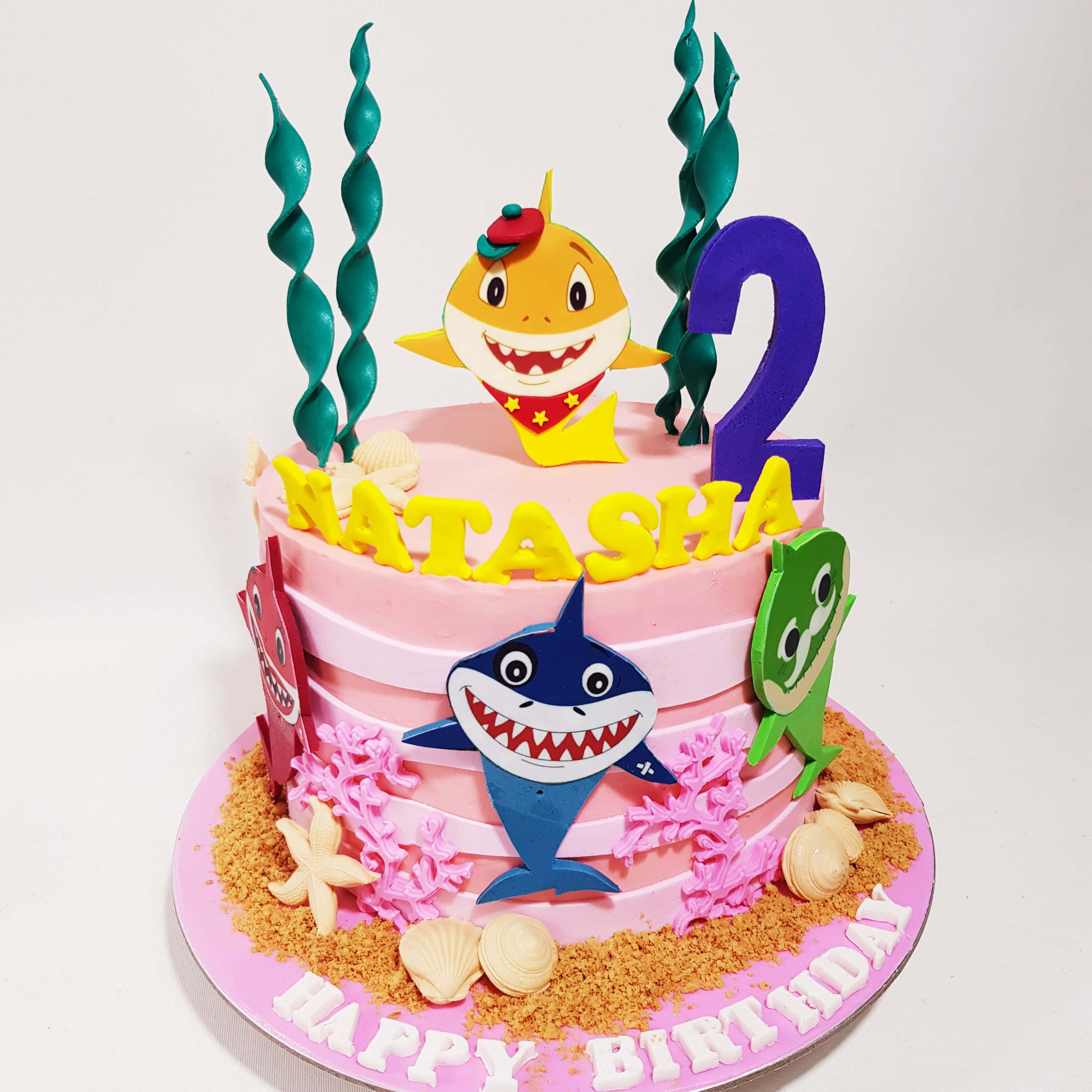baby shark 桌布 壁紙 — 一個粉紅色生日蛋糕，裝飾有Baby Shark角色、珊瑚貝殼和一個大數字二，以慶祝Natasha的兩歲生日。