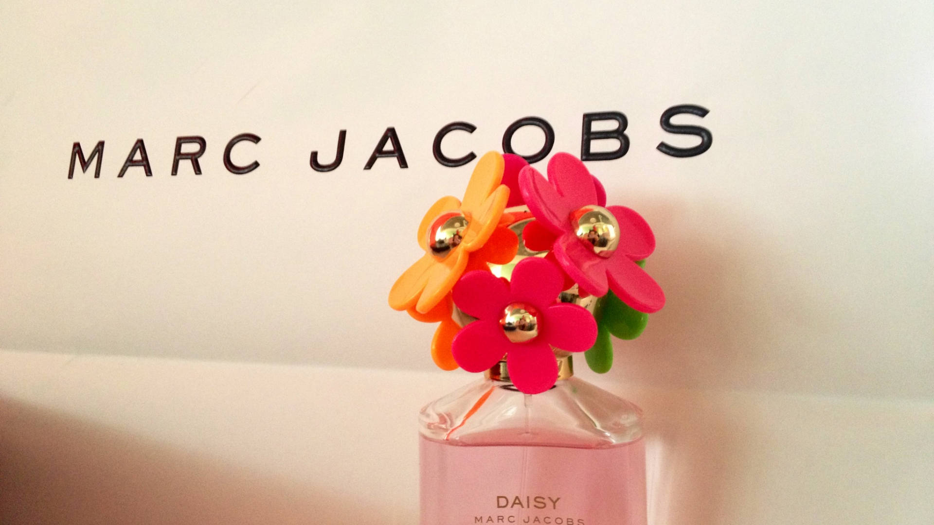 Marc Jacobs 桌布 壁紙 — 一個粉紅色的 Marc Jacobs Daisy 香水瓶，頂部飾有鮮橙色和粉紅色的塑膠花朵，靠著一面印有黑色品牌名稱的白牆。
