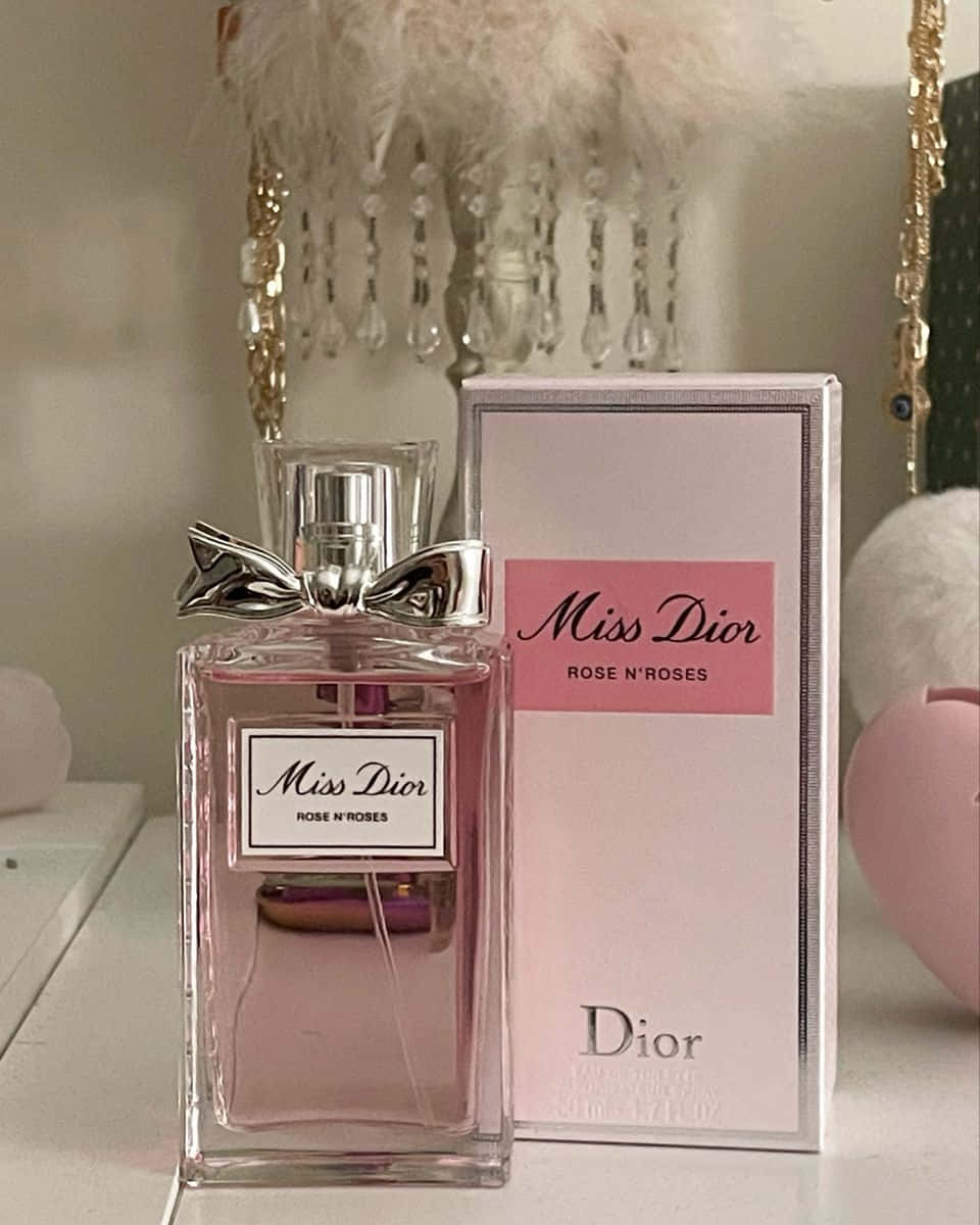 Rose Oil 桌布 壁紙 — 一個粉紅色的 Miss Dior 香水瓶，帶有銀色蝴蝶結，放在白色架子上，旁邊是它的盒子，還有 fluffy 粉紅色裝飾。
