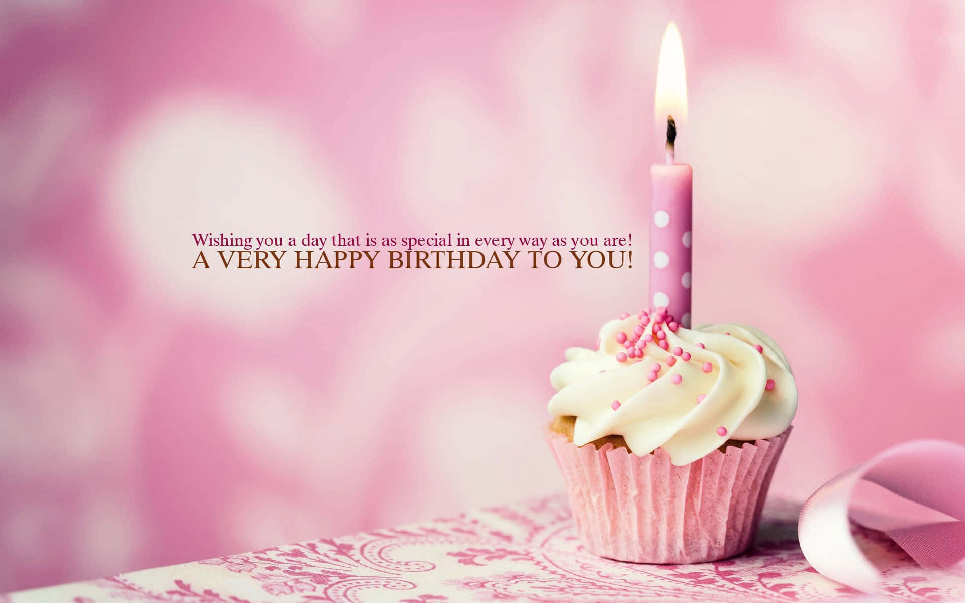 Birthday Wishes 桌布 壁紙 — 一個粉紅色的紙杯蛋糕，上面有白色糖霜和一支點燃的蠟燭，放在一個有圖案的表面上，背景是柔和的粉紅色心形散景。