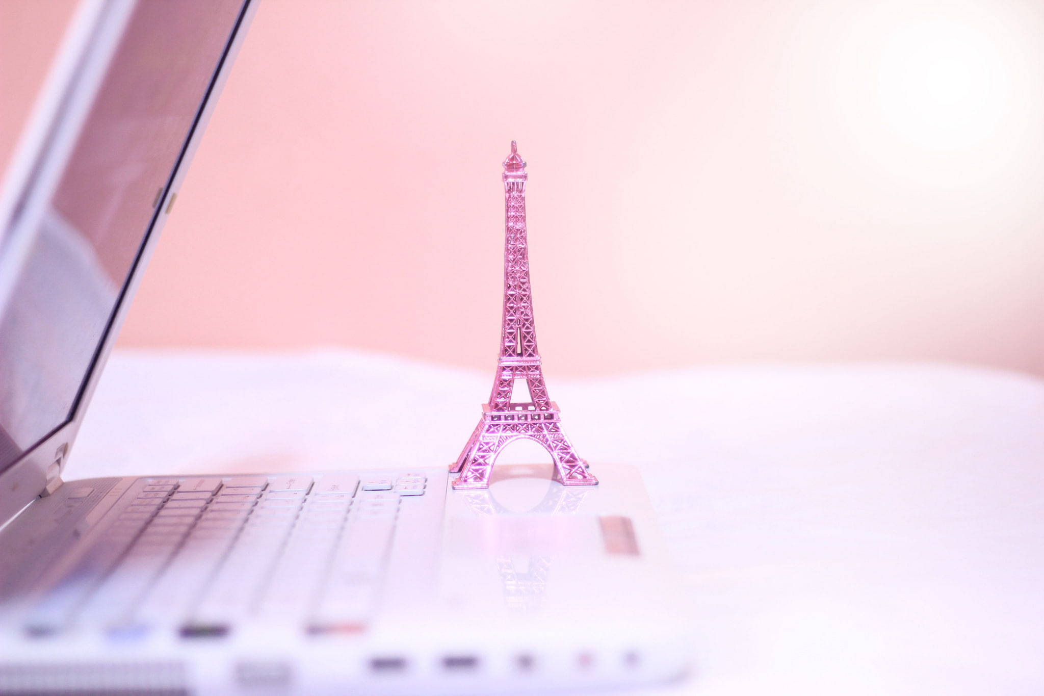 Eiffel Tower 桌布 壁紙 — 一個粉紅色的艾菲爾鐵塔小雕像優雅地立在白色筆記型電腦鍵盤上，背景是柔和的粉紅色。