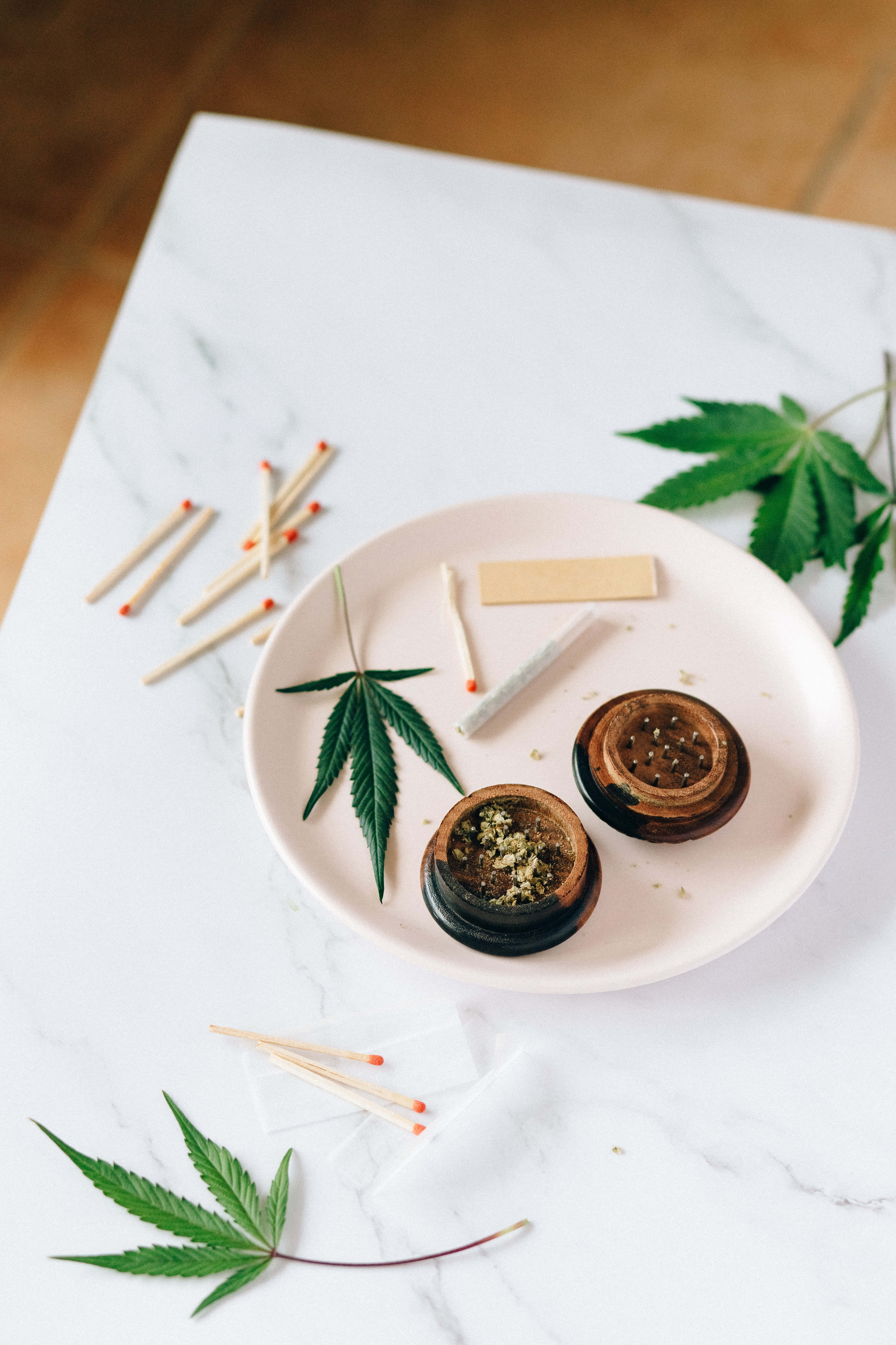 Weed Blunt 桌布 壁紙 — 一個粉紅色盤子上放著一個用過的大麻研磨器和捲菸紙，周圍是新鮮的綠葉和散落的火柴，置於大理石表面上。