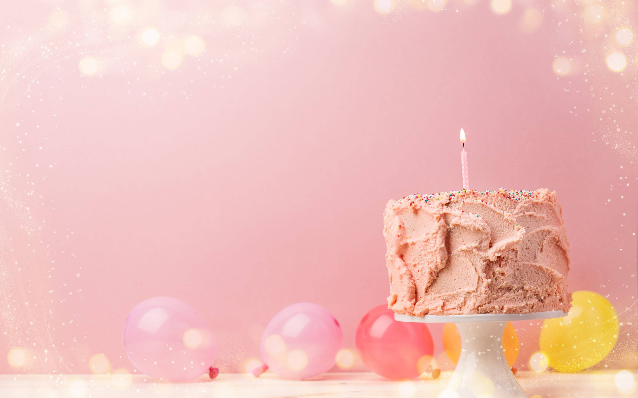 Birthday Cake 桌布 壁紙 — 一個粉紅色糖霜生日蛋糕，上面插著一支蠟燭，放在一個被彩色氣球環繞的架子上，背景是柔和的粉紅色散景。
