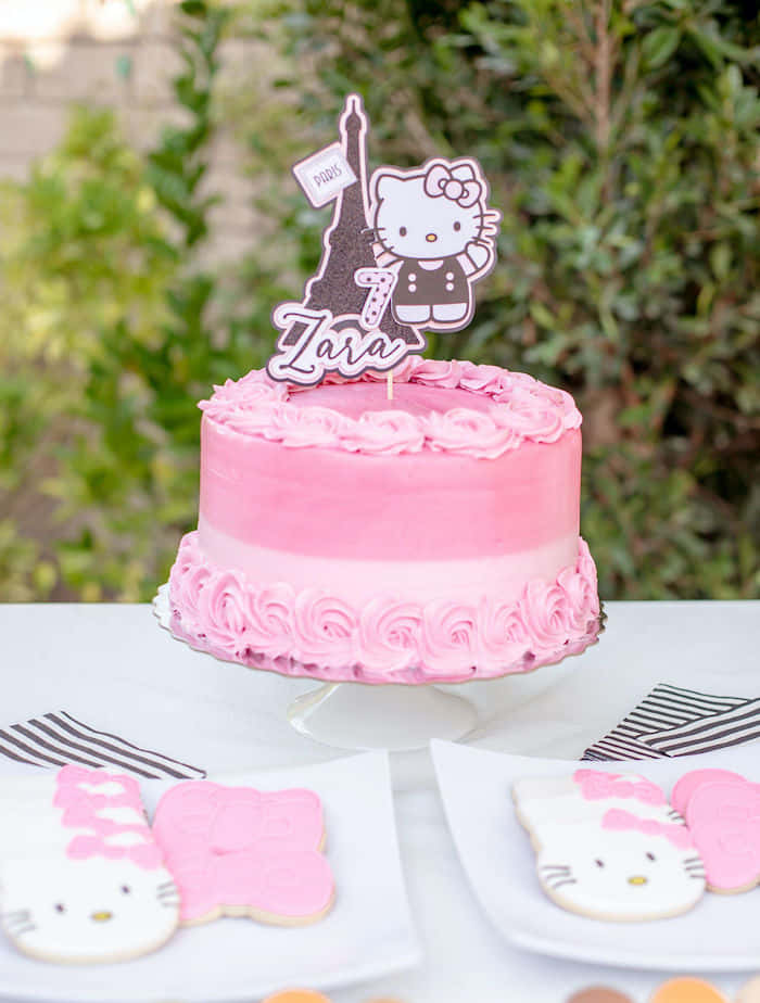 Hello Kitty Party 桌布 壁紙 — 一個粉紅色糖霜生日蛋糕，上面放著Hello Kitty和艾菲爾鐵塔裝飾，放在架子上，旁邊有主題餅乾。