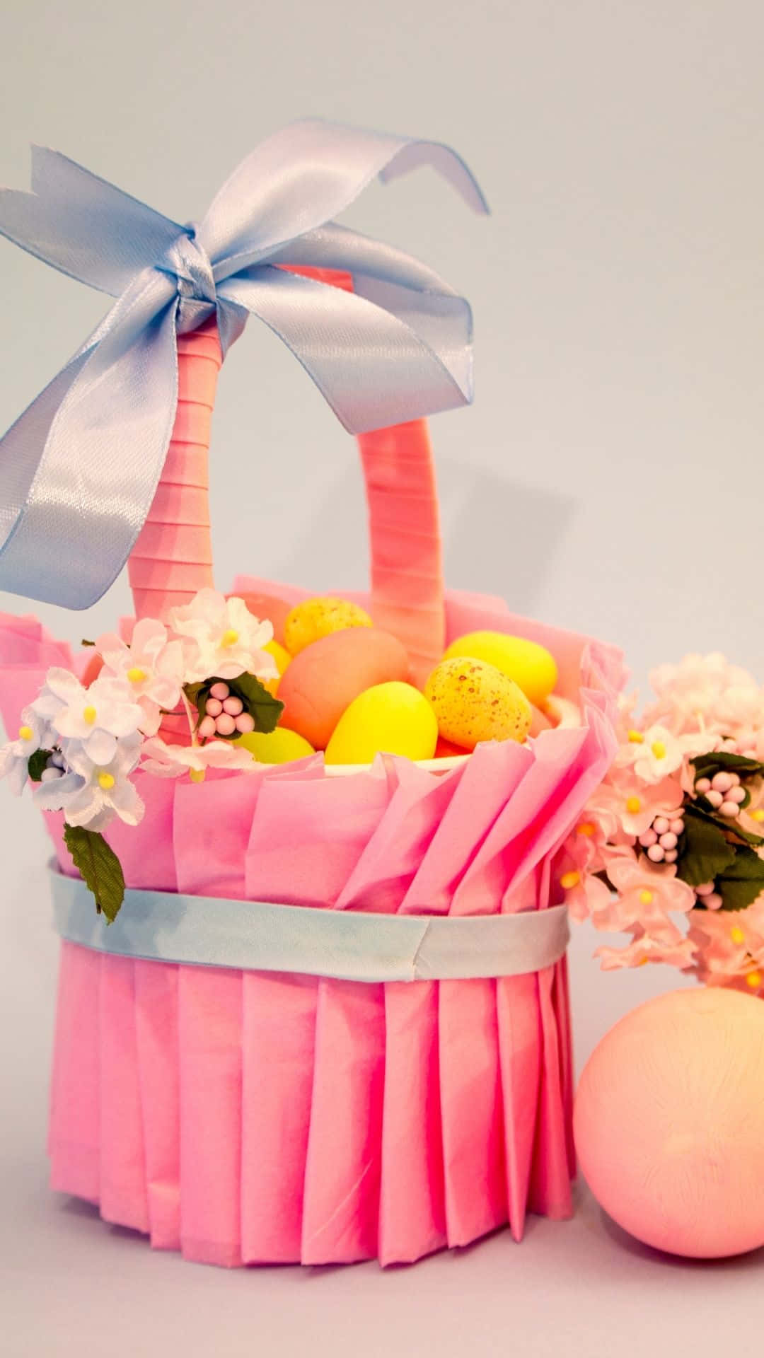 Easter Basket 桌布 壁紙 — 一個粉紅色紙籃，飾有淺藍色絲帶，裝著色彩繽紛的斑點蛋和白色花朵，背景是柔和的灰色。