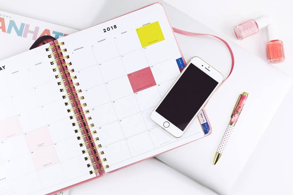 iPhone 桌布 壁紙 — 一個粉紅色螺旋計劃本，2018年日曆打開，旁邊放著智慧型手機、花卉筆和指甲油，在白色表面上。