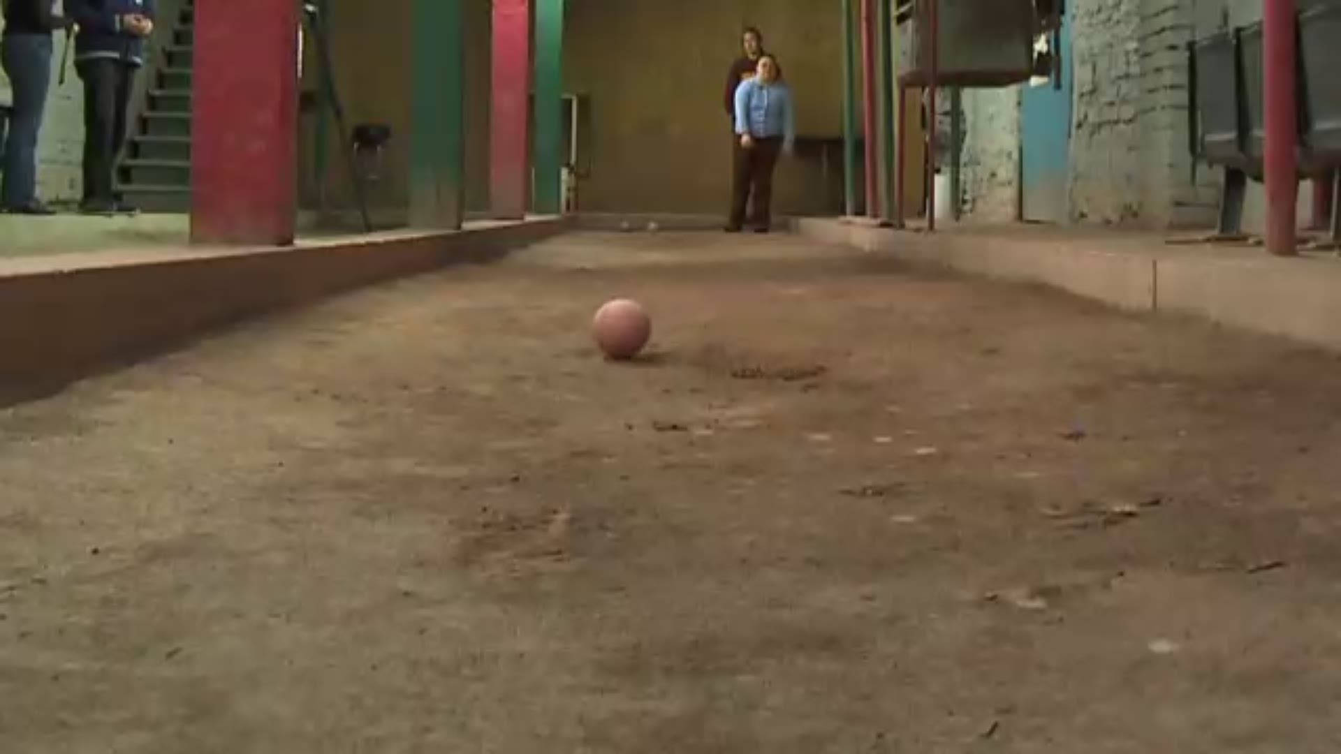 運動 桌布 壁紙 — 一個粉紅色足球放在狹窄庭院的塵土飛揚的地面上，附近有人站在色彩繽紛的柱子旁。