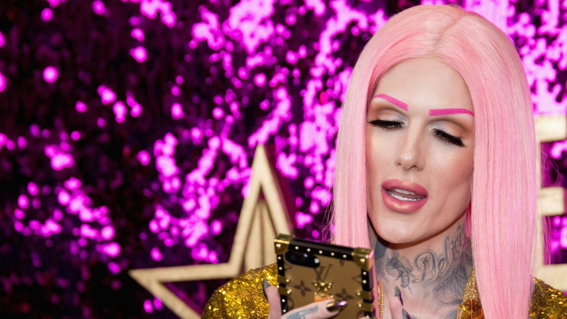 Jeffree Star 桌布 壁紙 — 一個粉紅色頭髮、化著大膽妝容的人，在閃爍的紫色背景前拿著一部金色手機。