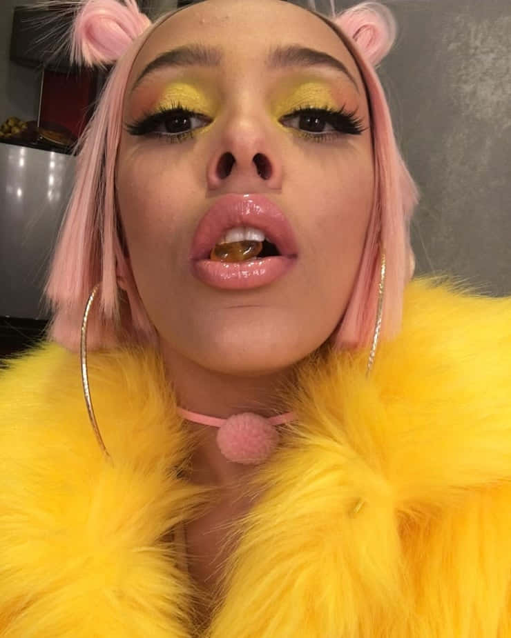 Doja Cat 桌布 壁紙 — 一個粉紅色頭髮、豬耳朵的人穿著鮮黃色毛皮大衣和黃色眼妝，做出調皮的表情。