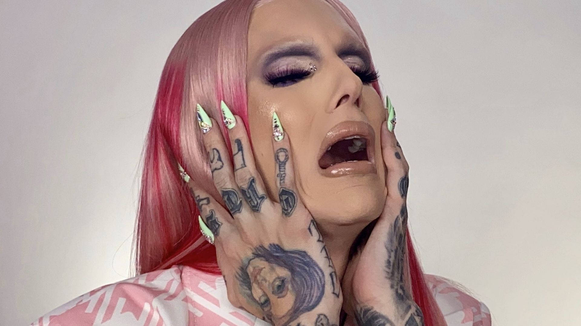Jeffree Star 桌布 壁紙 — 一個粉紅色頭髮和紋身雙手的人，在純色背景前以戲劇性的特寫將手掌壓在臉頰上。
